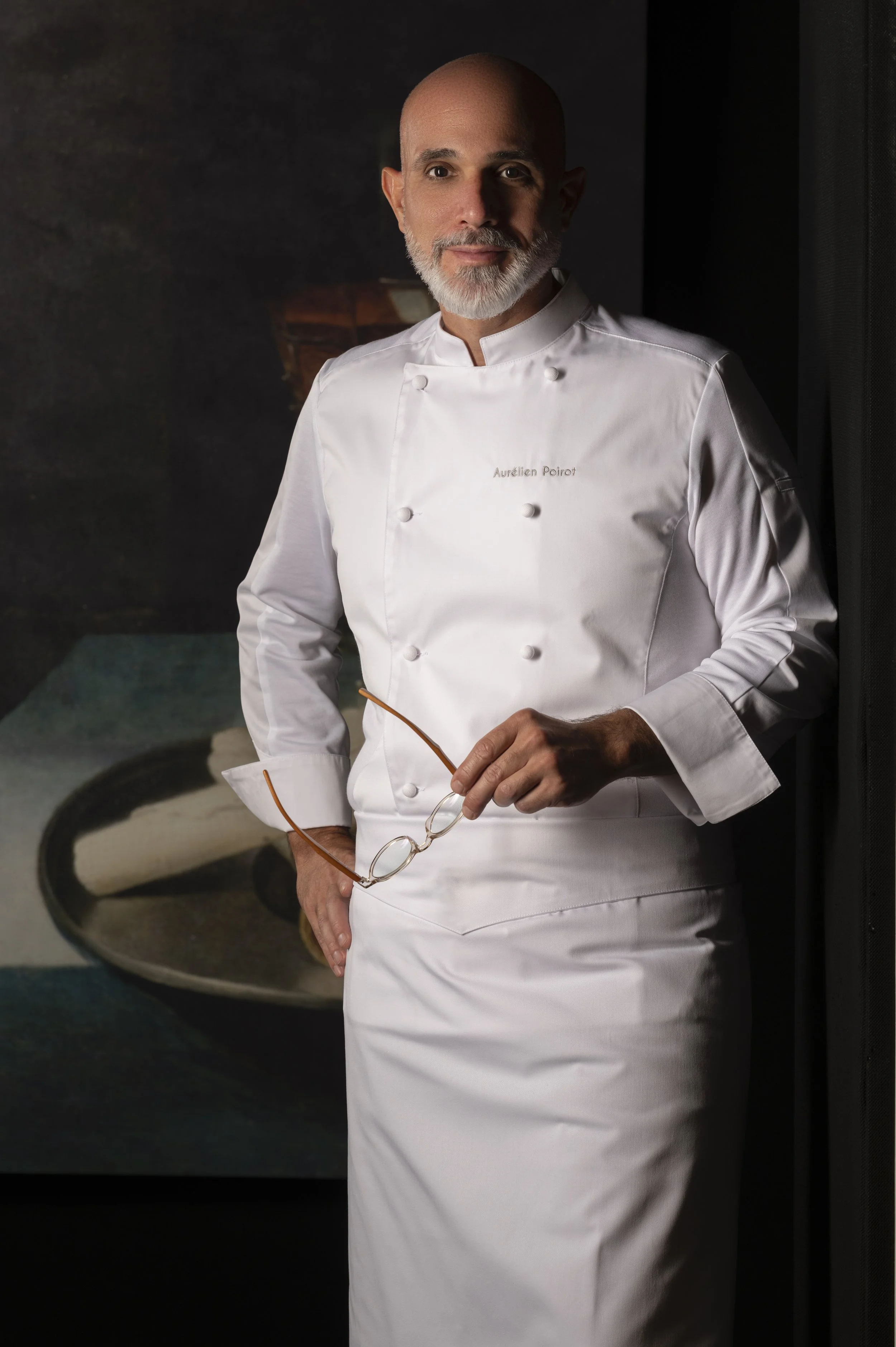 Un chef en tenue blanche, tenant une paire de lunettes, posant devant une peinture