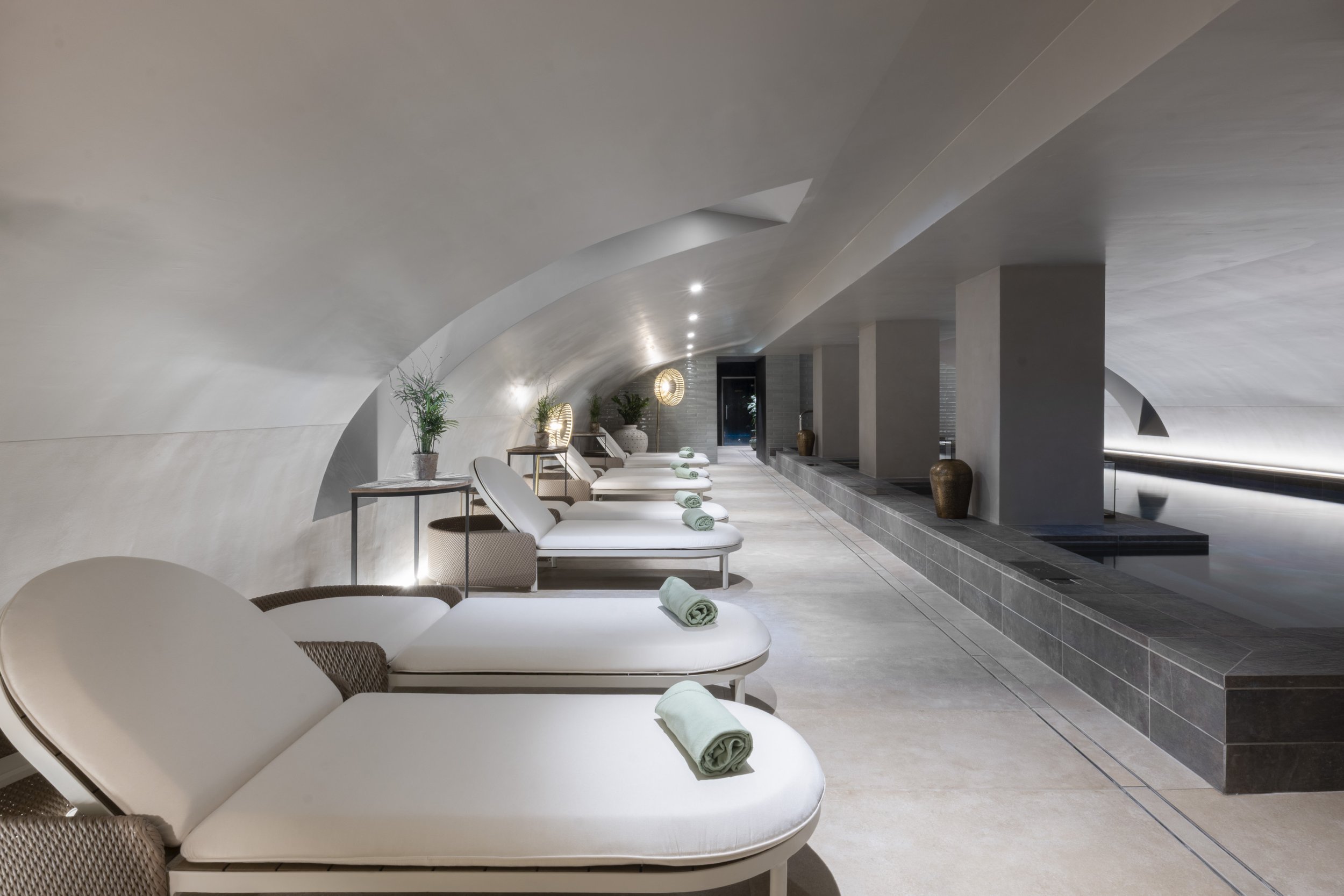 Série de transats blancs avec des serviettes vertes pliées, dans un espace spa moderne avec murs en courbe et décoration minimaliste.