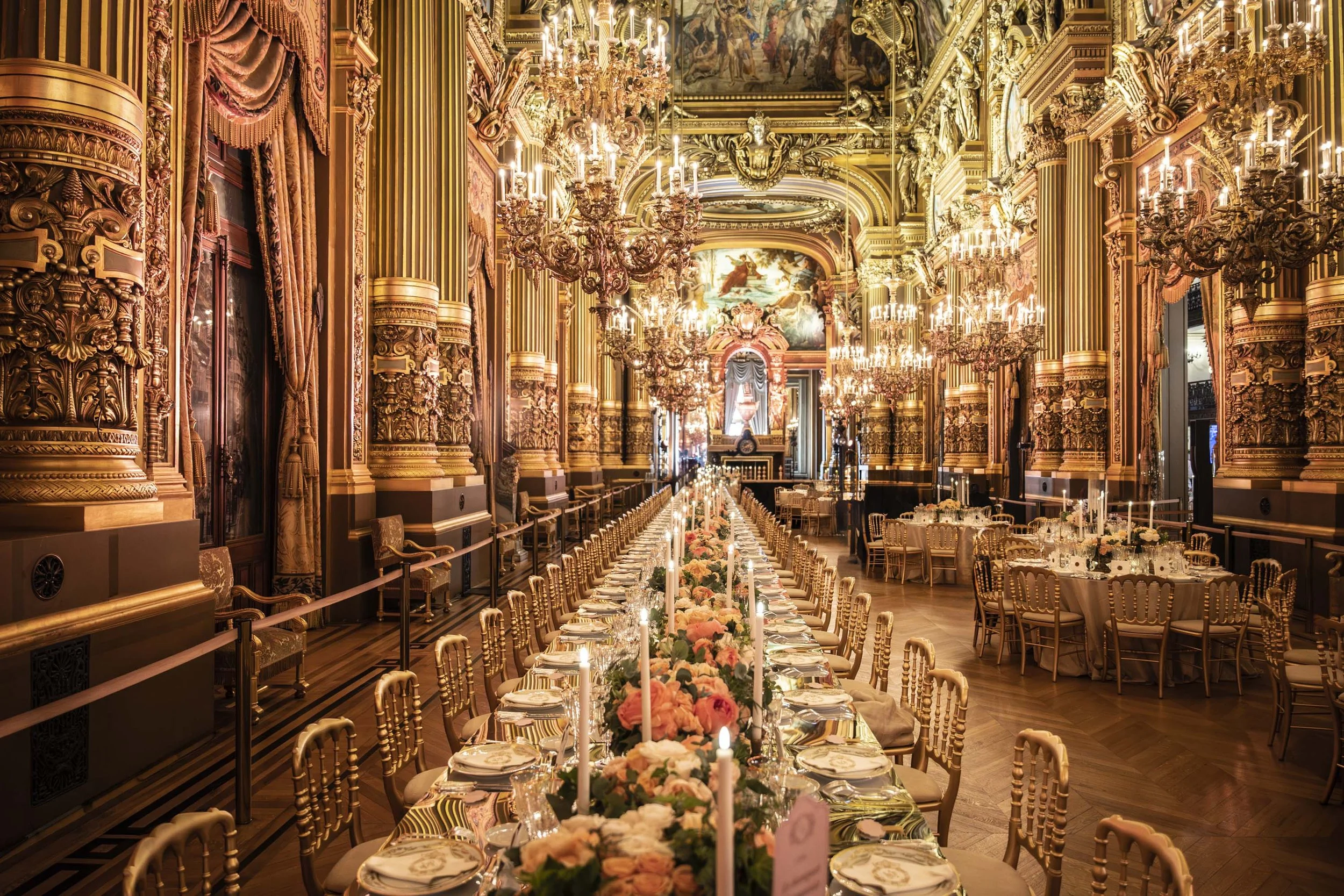 Une salle de banquet somptueuse avec un long table décoré de fleurs et de chandelles, entourée de chaises dorées dans une pièce richement ornée de moulures dorées, de rideaux en velours, de chandeliers en cristal suspendus et d'œuvres d'art au plafon