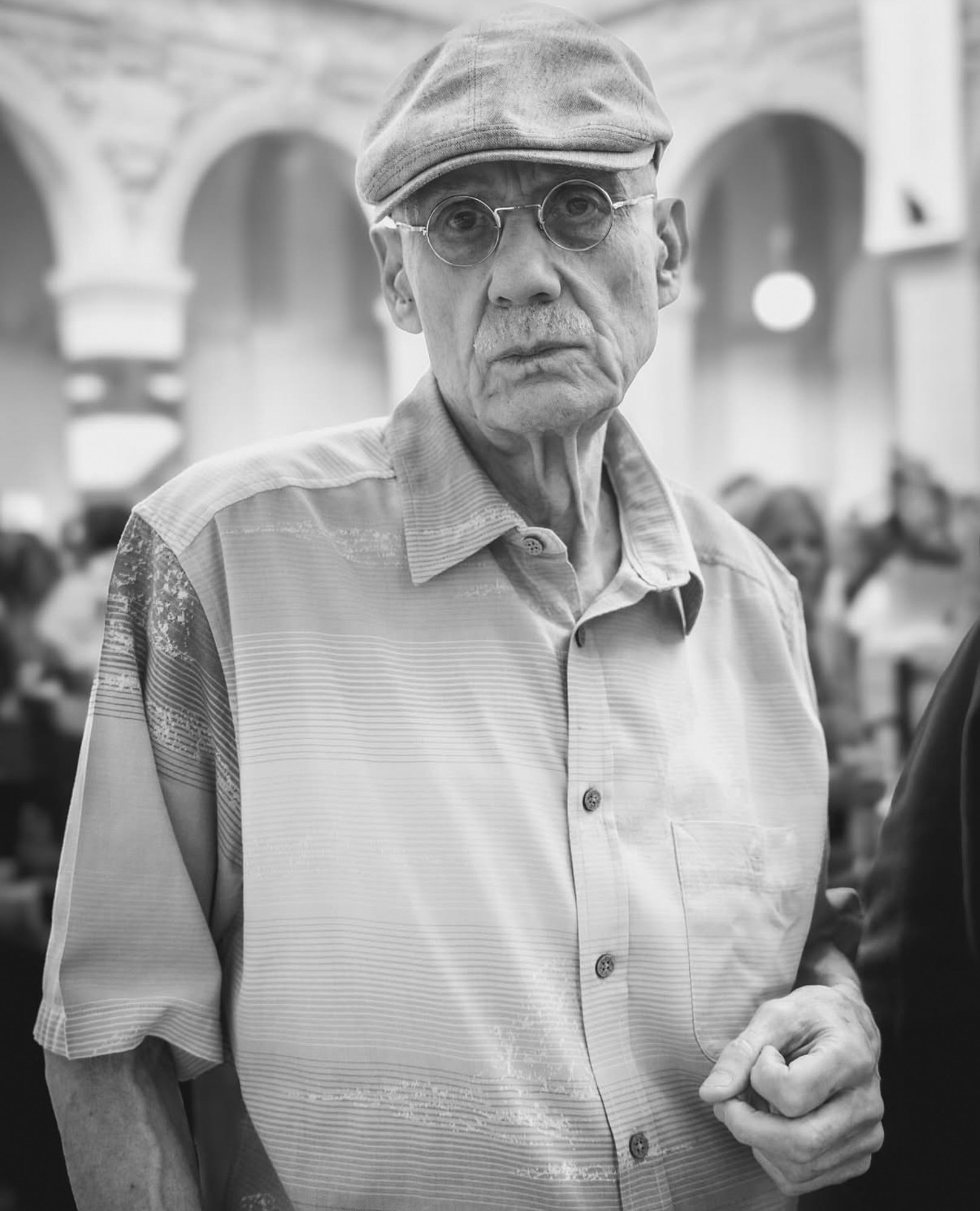 Un vieil homme portant une chemise à rayures et un bonnet, portant des lunettes rondes, dans un espace intérieur, peut-être un marché ou une place publique.