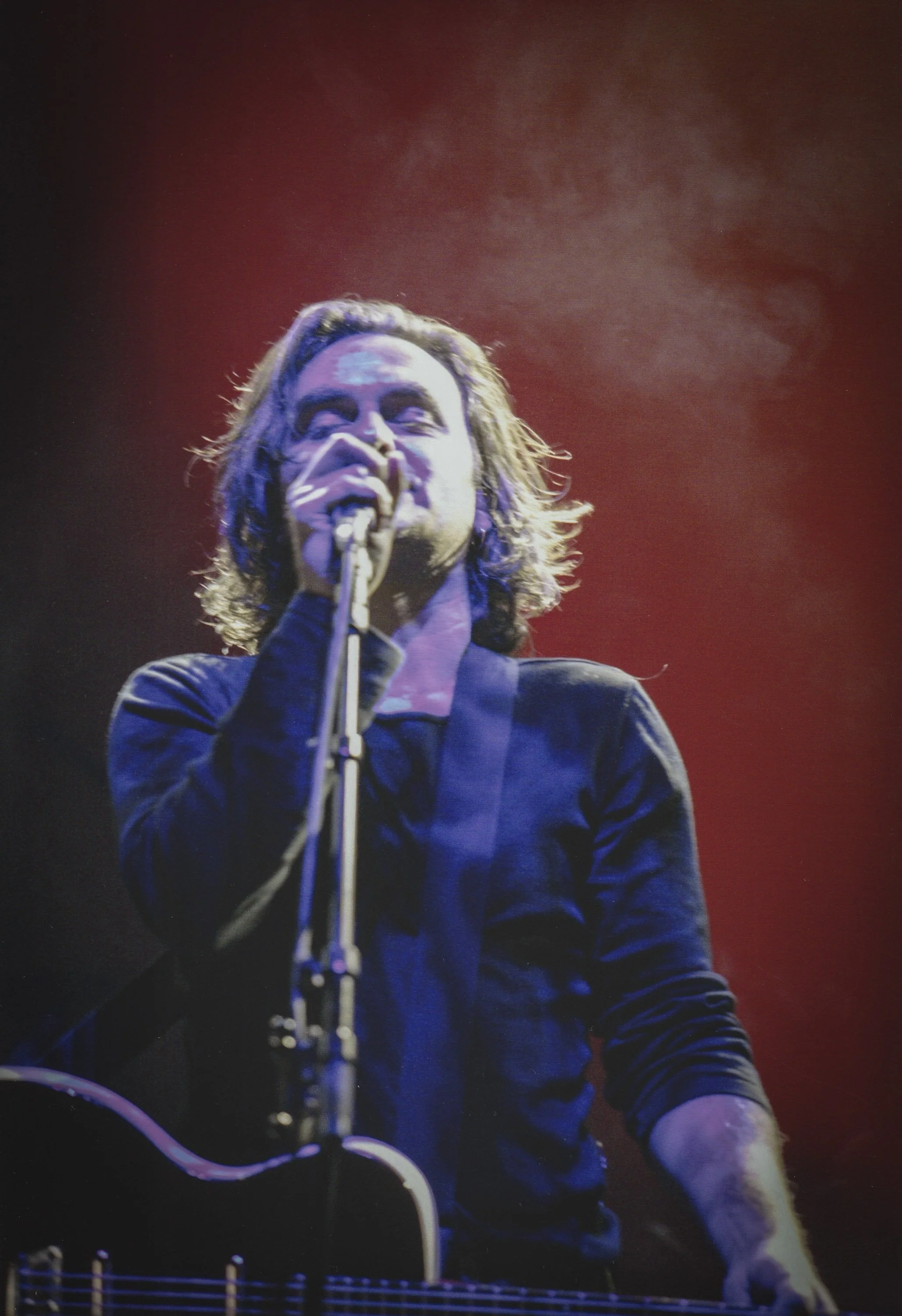 Un homme chante avec un micro, tenant une guitare, sur scène avec un fond rougeâtre.