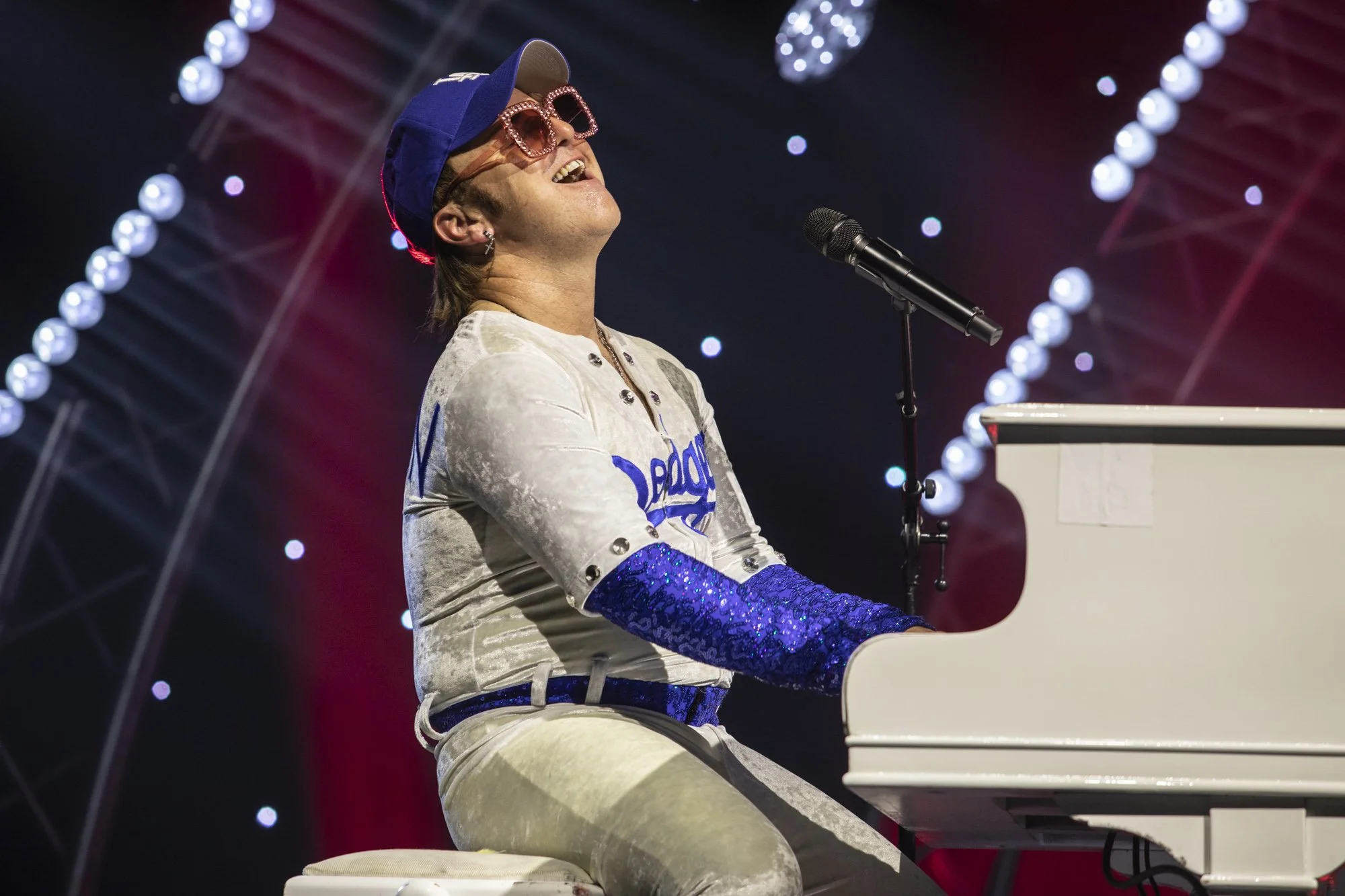 Une personne joue du piano sur scène, portant des vêtements brillants, un chapeau et de grosses lunettes à cadre rose, souriant joyeusement.