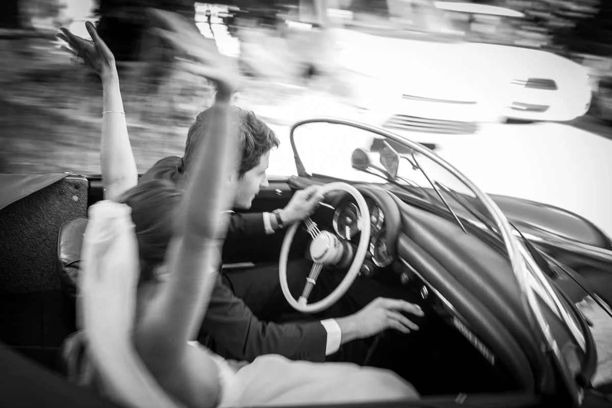 Deux personnes dans une voiture de course ancienne, le conducteur au volant et un passager levant la main, en mouvement rapide, en noir et blanc.