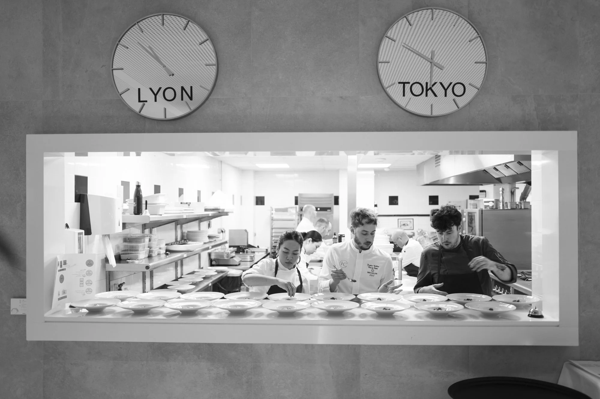Vue en noir et blanc de la cuisine d'un restaurant vue à travers une fenêtre, avec des chefs et cuisiniers préparant des plats, deux horloges au-dessus affichant l'heure à Lyon et Tokyo.