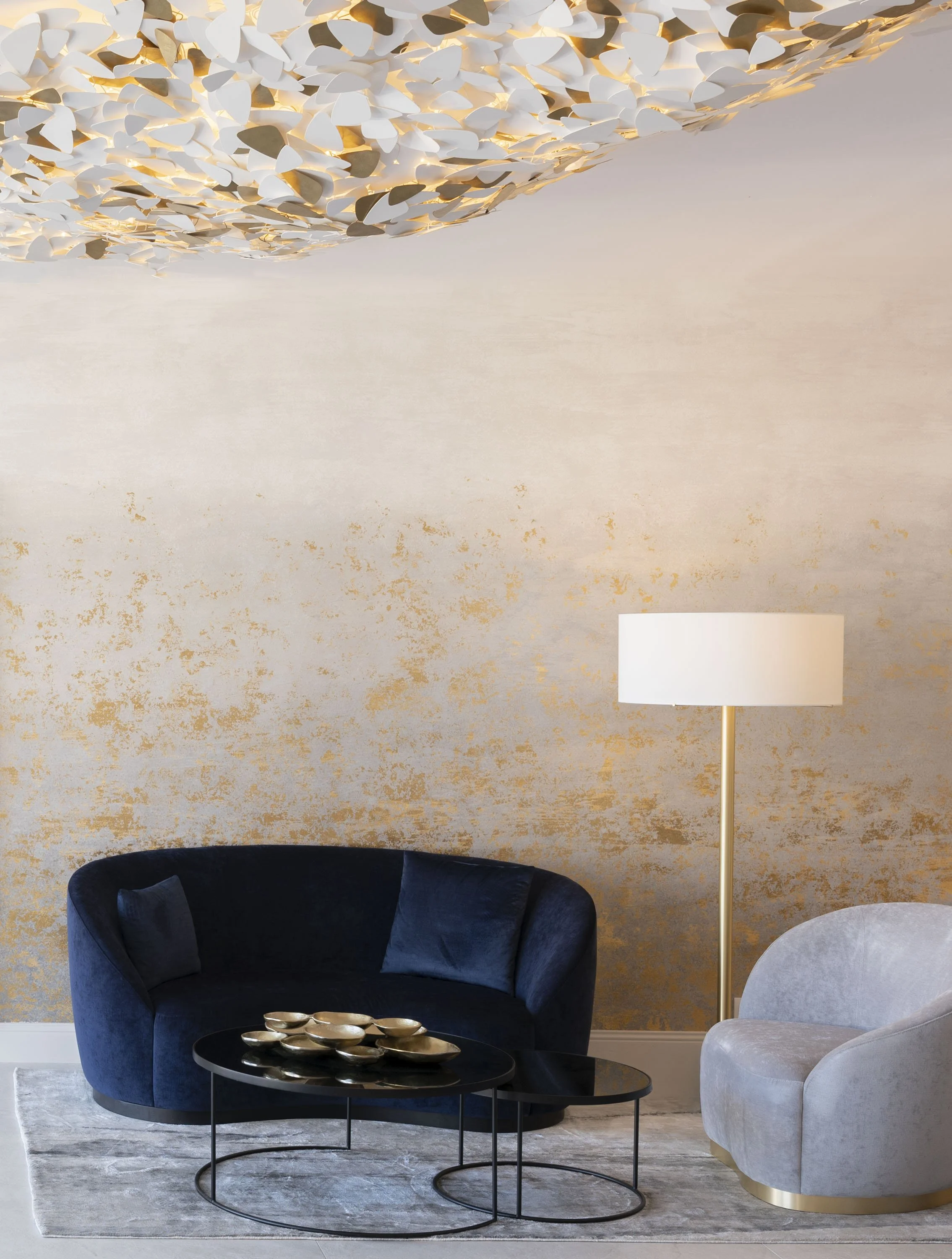 Salon moderne avec un canapé bleu foncé, un fauteuil gris clair, une lampe de sol, deux tables basses noires et une décoration murale dorée