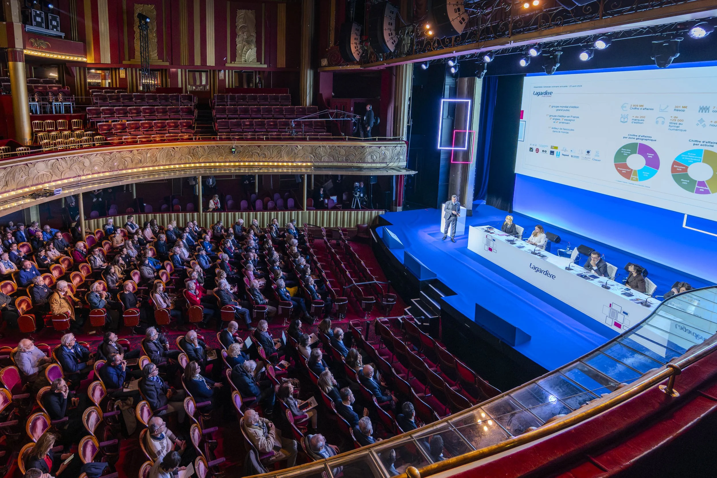 Salle de conférence avec un public assis, un présentateur sur scène et un panneau d'affichage numérique