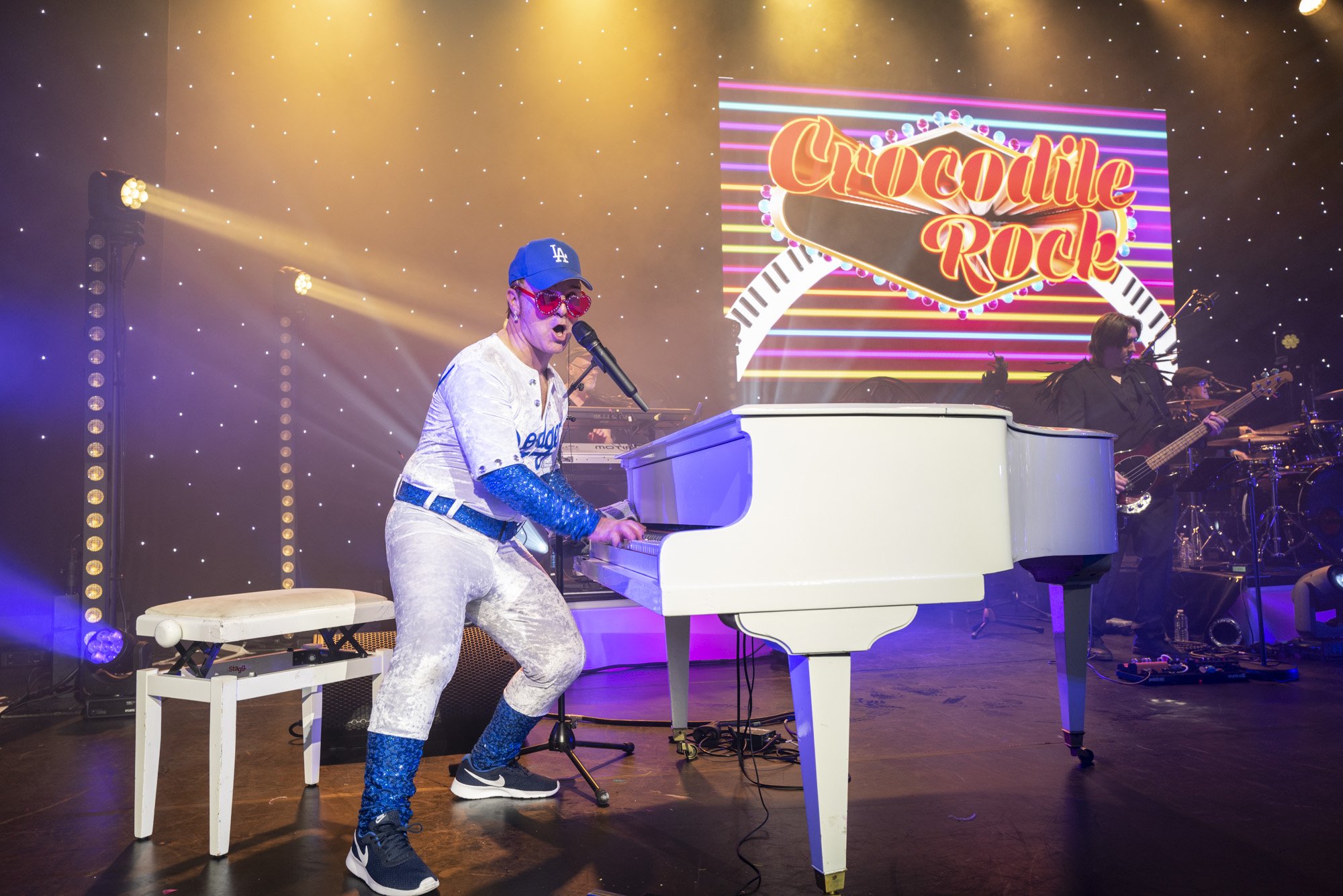 Une performance musicale avec un homme jouant du piano blanc sur scène, portant une tenue argentée et des lunettes de soleil roses, avec un groupe en arrière-plan et un écran lumineux affichant 'Crocodile Rock'.