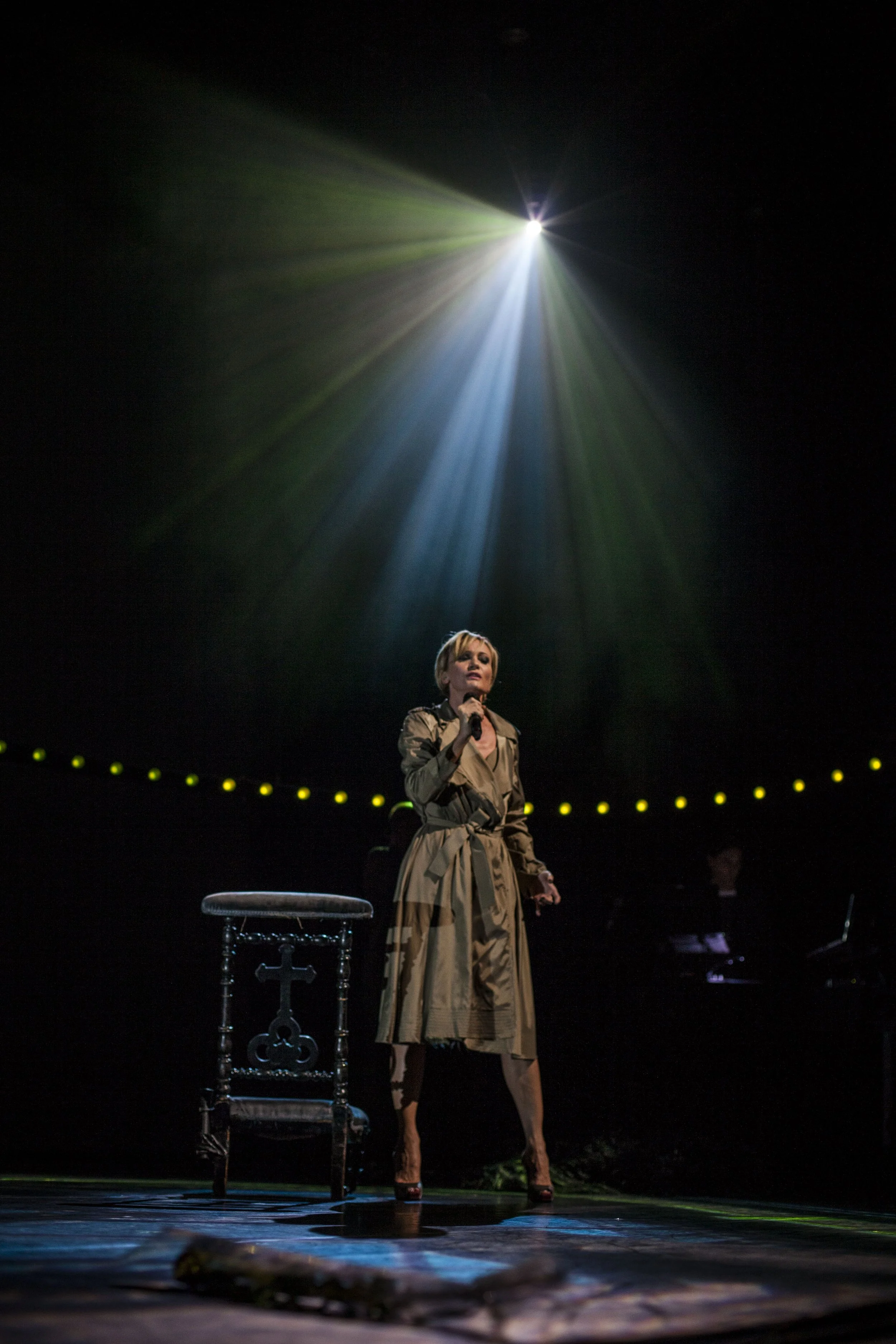 Une femme chante sur scène sous un éclairage dramatique, avec un microphone à la main, un siège en bois à côté d'elle.