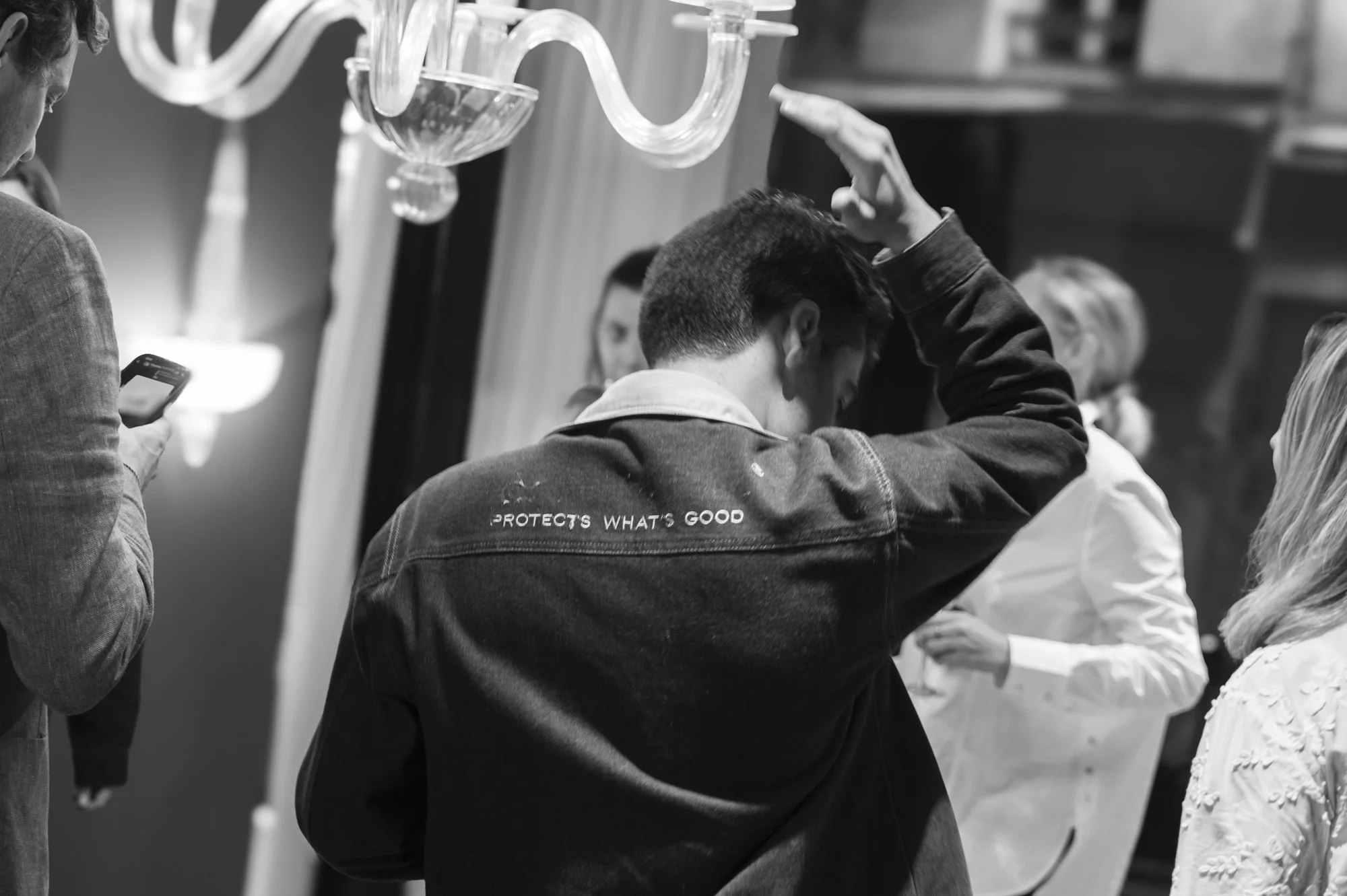 Groupe de jeunes adultes en conversation dans un lieu intérieur, certains utilisant leur téléphone. Noir et blanc.