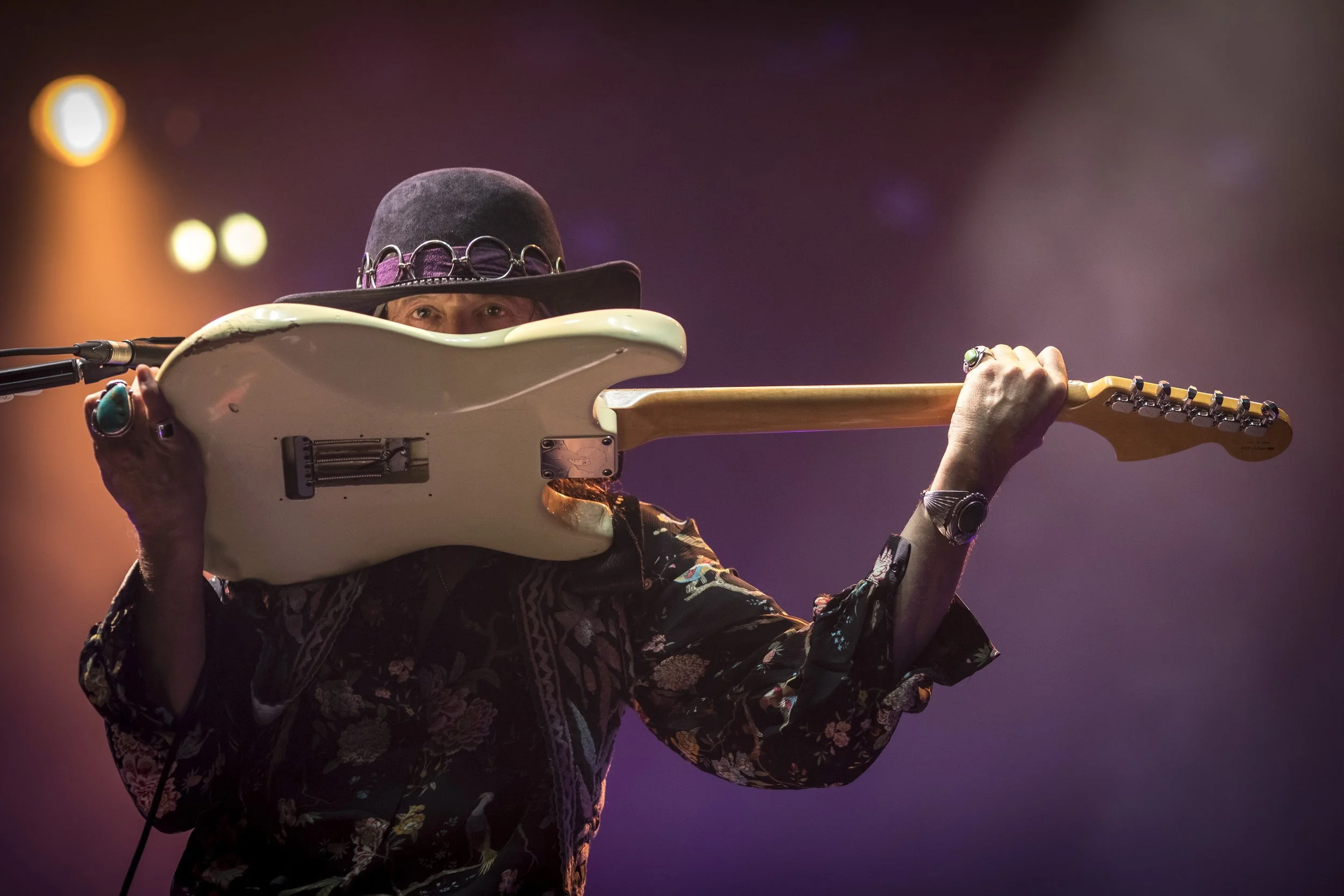 Musicien portant un chapeau noir et jouant de la guitare électrique blanche lors d'une performance sur scène, avec un éclairage chaud en arrière-plan.