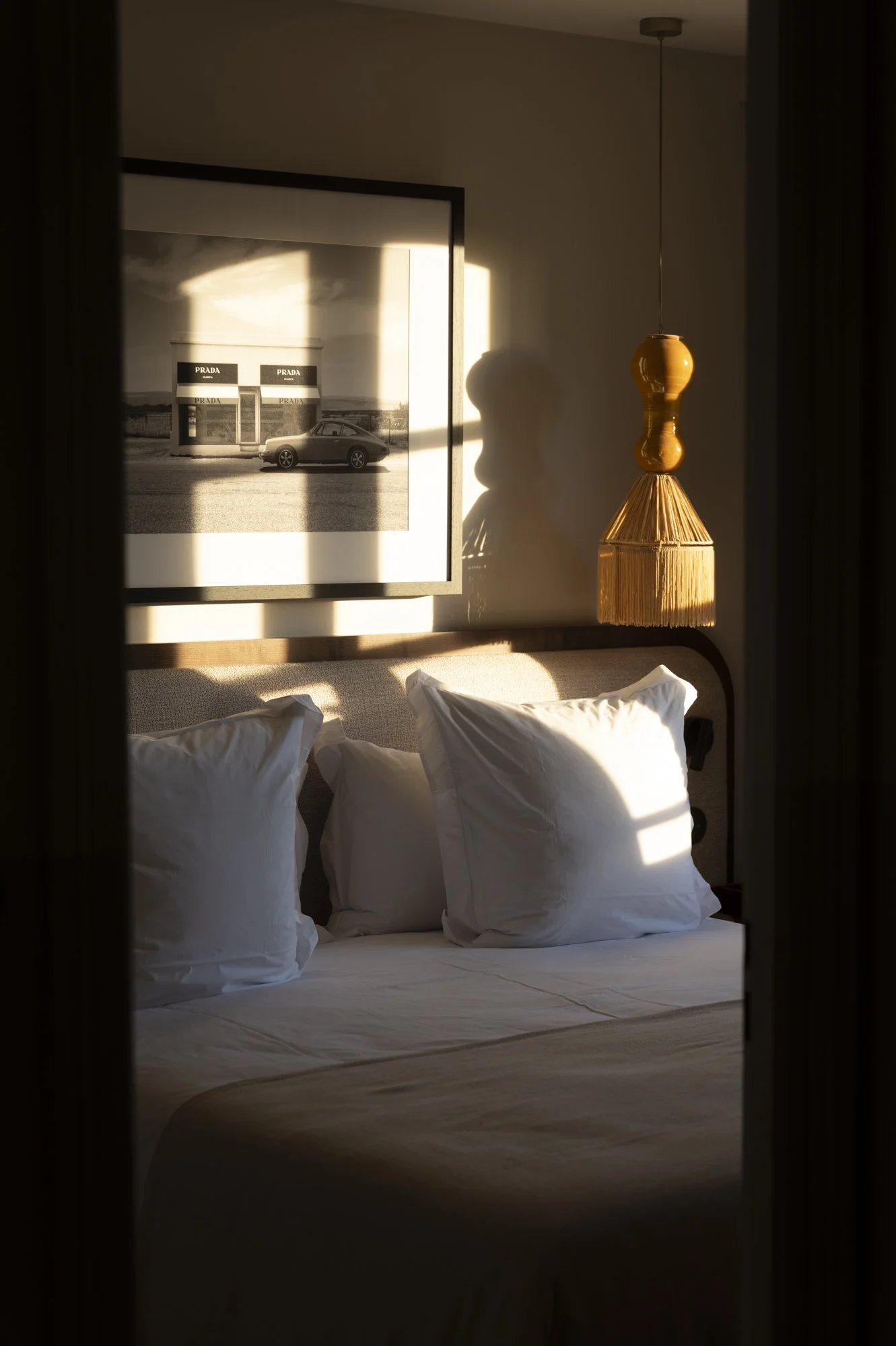 Un lit avec des oreillers blancs dans une chambre, éclairé par la lumière du soleil, avec un tableau noir et blanc d'une voiture et d'une boutique Prada, et un luminaire ornemental en bois suspendu au plafond.