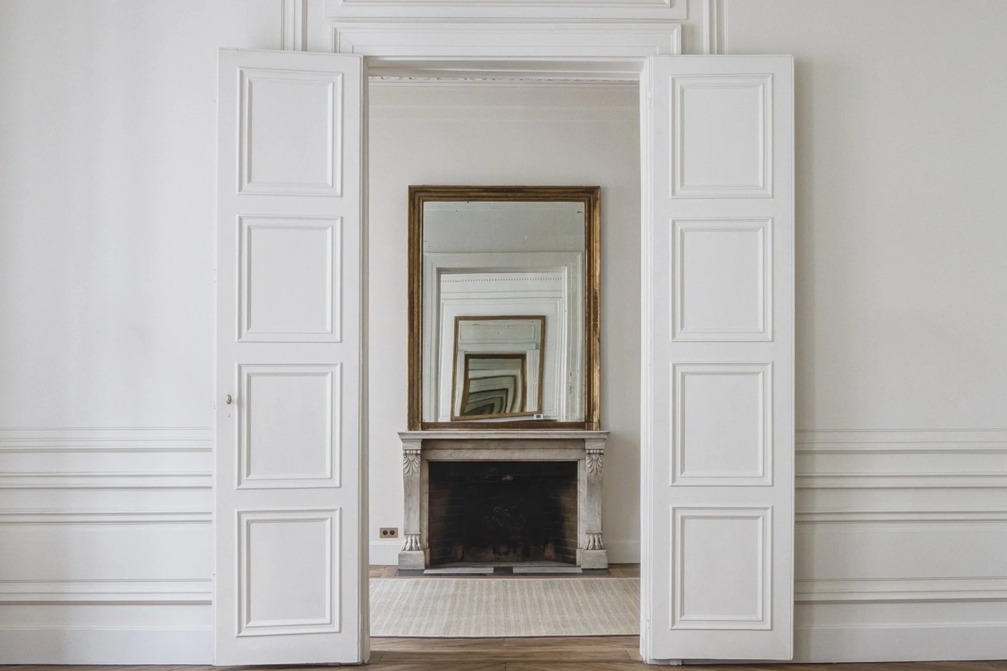 Salle avec cheminée en marbre, grand miroir encadré de bois doré, murs blancs avec moulures, plancher en bois, porte en bois peint en blanc.