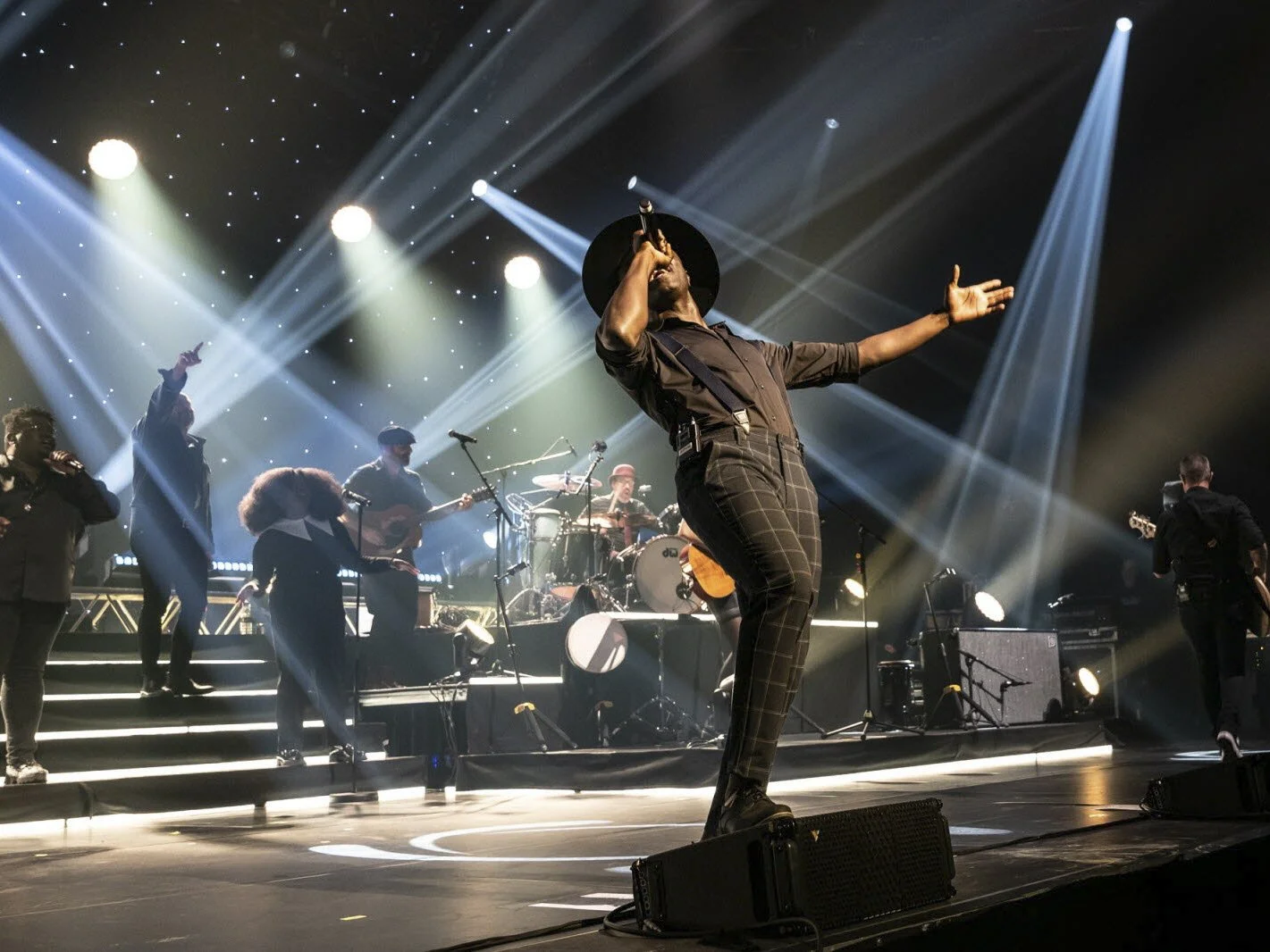 Un groupe de musiciens en concert avec un chanteur au centre, tenant un microphone, sous un éclairage de scène dramatique et des lumières colorées.