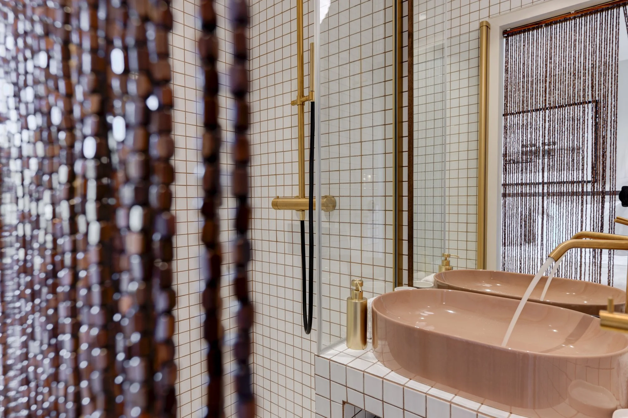 Salle de bain avec deux lavabos roses, miroir, distributeur de savon doré, et rideau en perles.