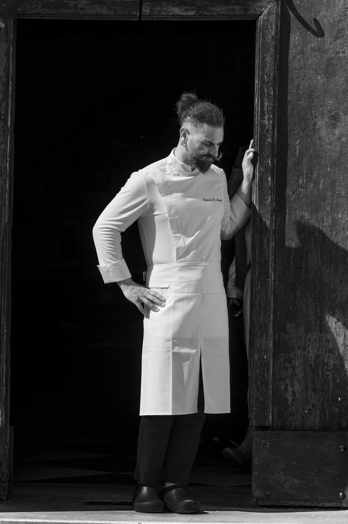 Un chef en tenue blanche se tient dans l'entrée d'une cuisine, semblant réfléchir ou en pause, en noir et blanc.