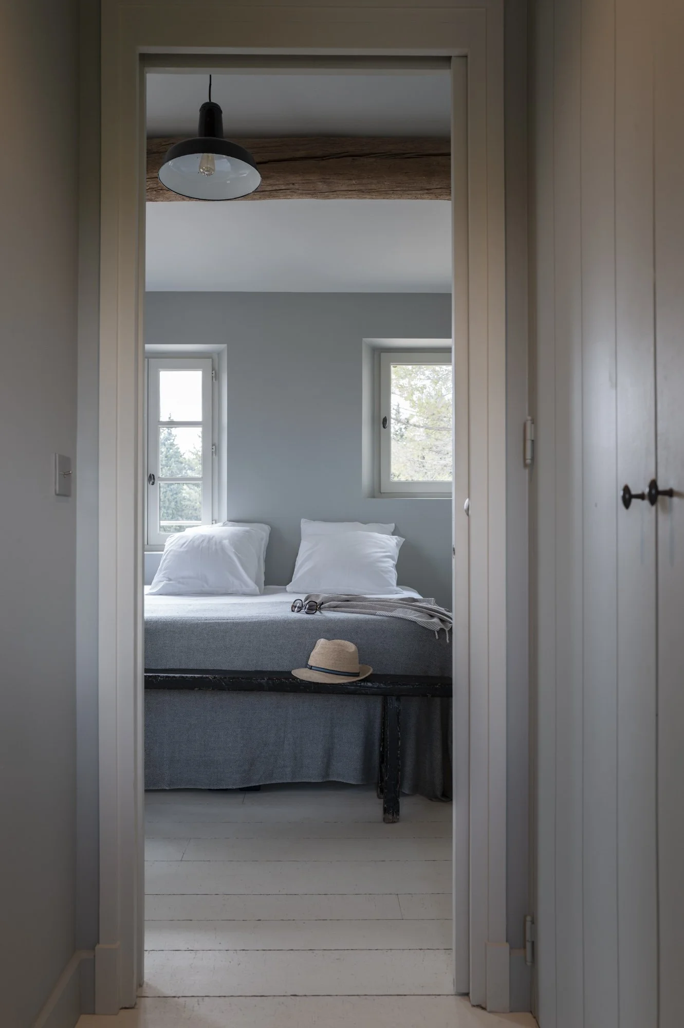 Une chambre confortable avec deux fenêtres, un lit avec des oreillers blancs, une paire de lunettes, un chapeau en paille et une couverture sur le lit, vue depuis un couloir.