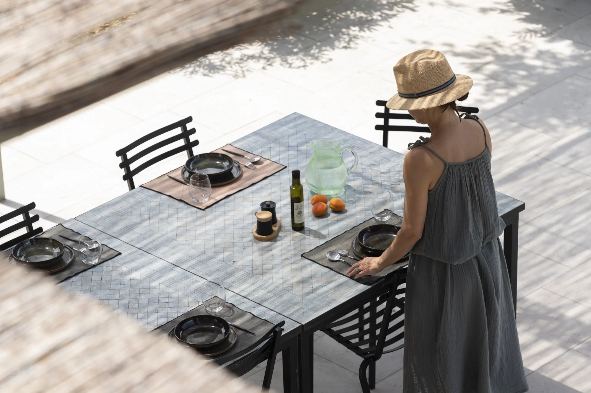 Une femme portant un chapeau en paille, une robe grise, prépare la table de repas en extérieur avec des assiettes, verres, une carafe d'eau, une bouteille d'huile d'olive, et des fruits sur la table.