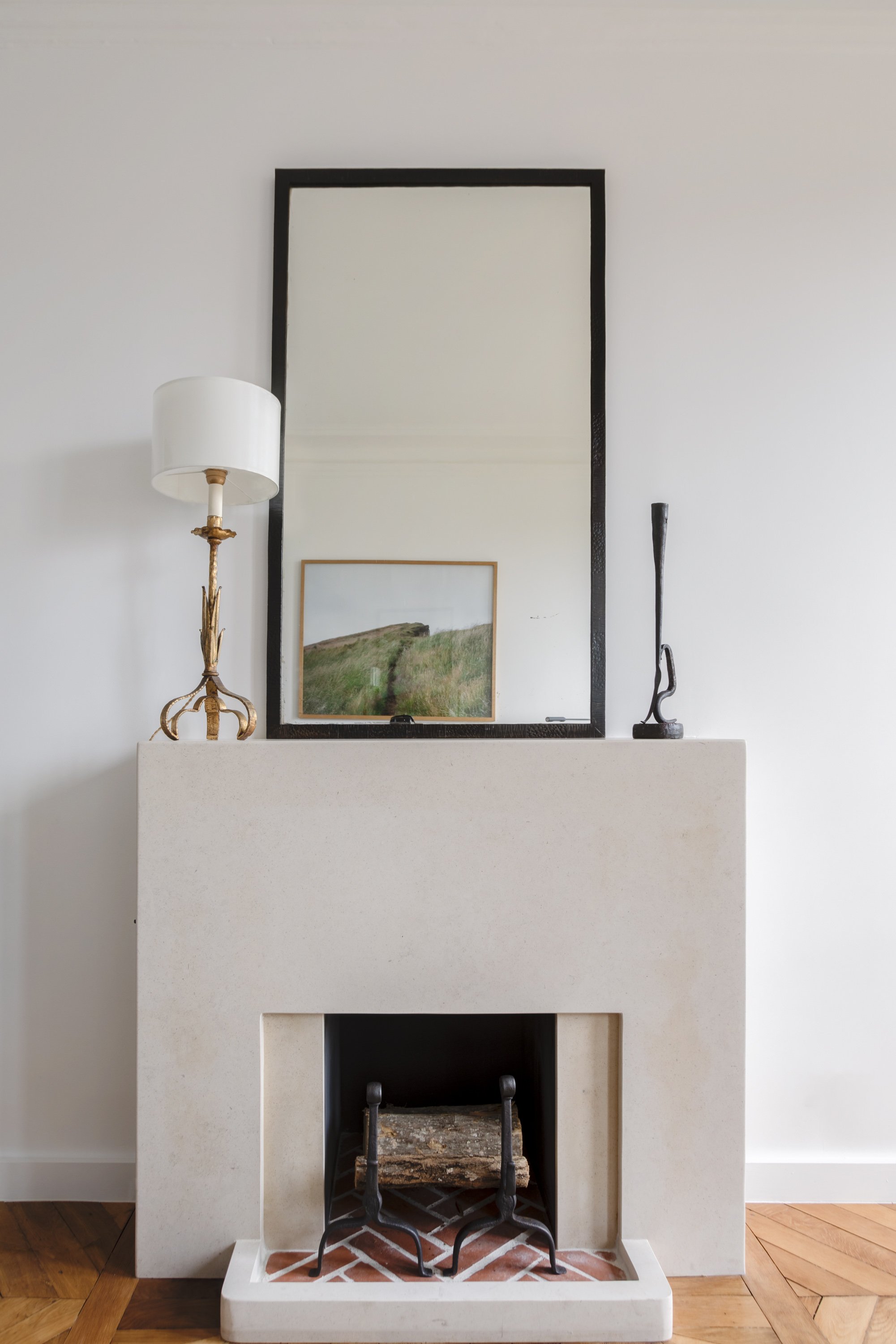 Un cheminée en pierre blanche avec un miroir rectangulaire noir au-dessus. Sur le manteau, il y a une lampe de table avec un abat-jour blanc à gauche et une sculpture noire à droite. Le feu est cassé, avec du bois dans la cheminée.