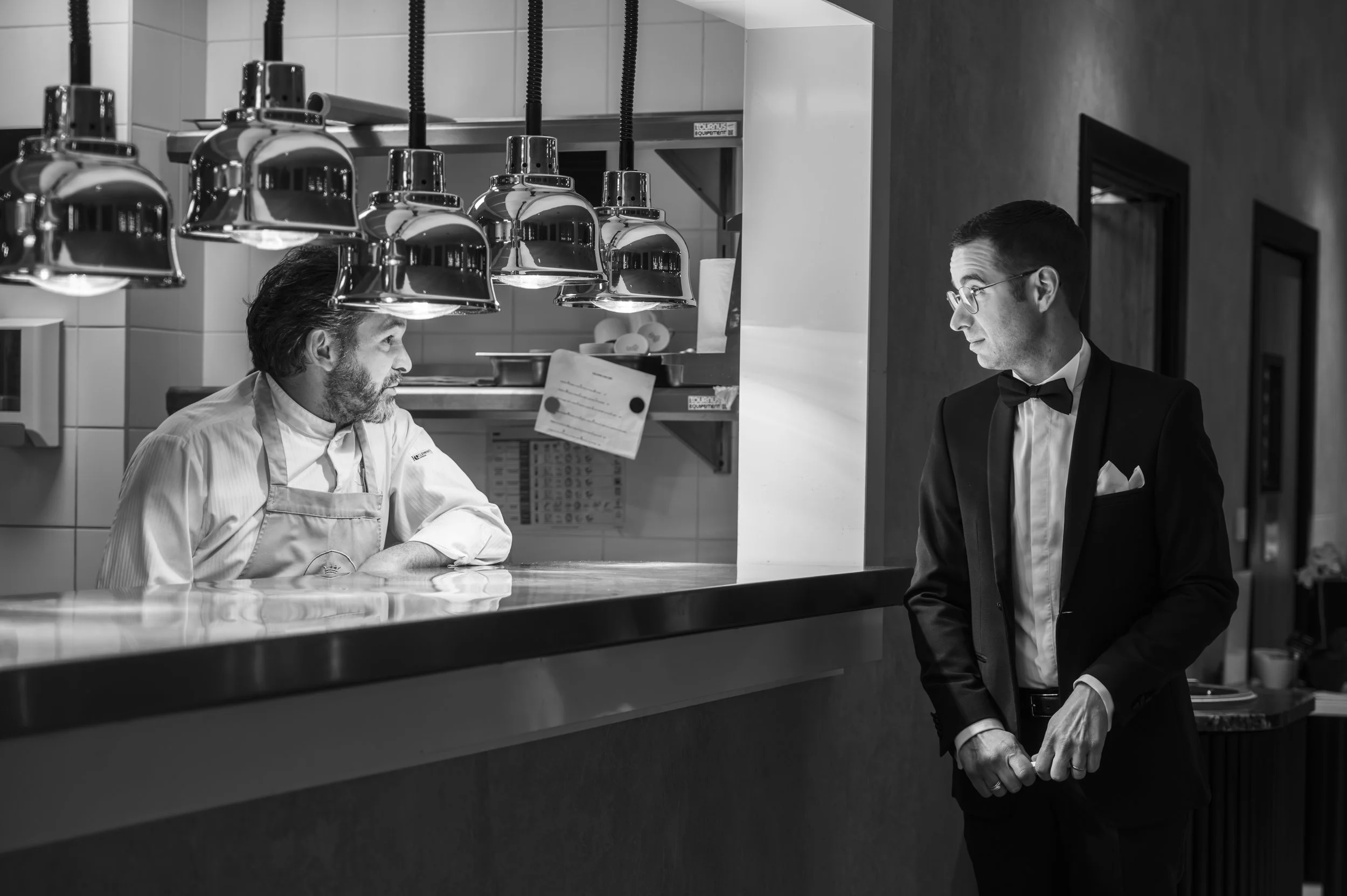 Un homme en costume noir avec une chemise blanche et une pochette, portant un nœud papillon, se tient devant un comptoir de restaurant, discutant avec un chef en uniforme blanc derrière le comptoir en cuisine.