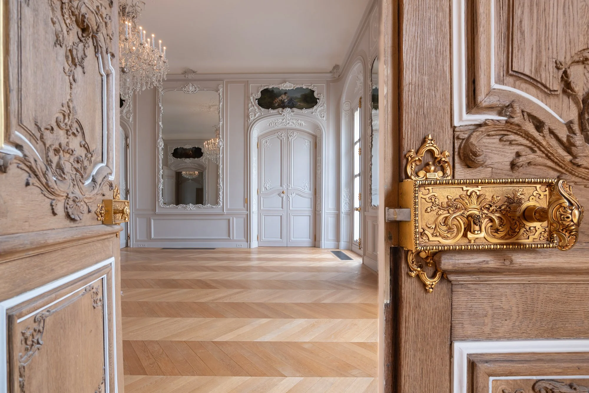 Une salle élégante avec des murs blancs ornés de moulures, un chandelier en cristal, des miroirs avec des cadres ornés, un parquet en bois clair, une porte blanche avec des détails sculptés, et une porte en bois avec une poignée dorée décorative.