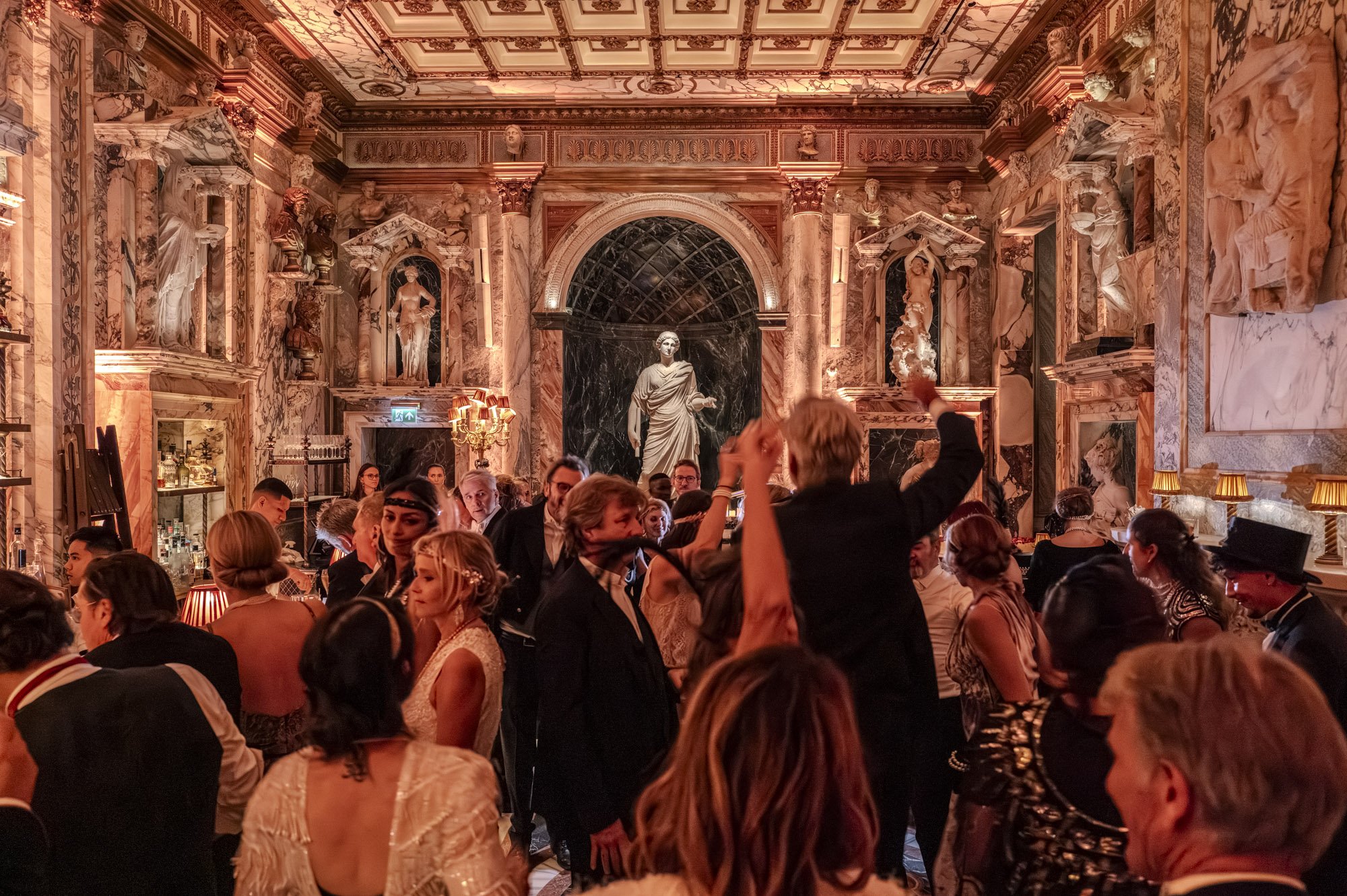 Une salle de réception élégante et luxueuse, décorée dans un style classique avec des sculptures et des fresques au plafond. Les invités, habillés en tenues formelles, dansent et socialisent dans une atmosphère festive.