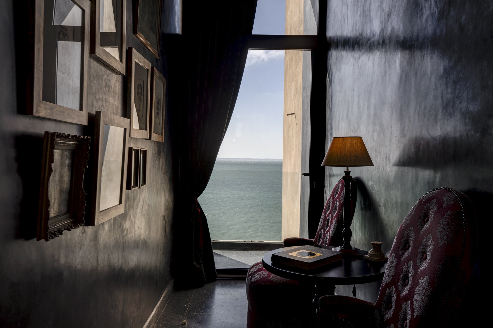 Intérieur sombre d'une pièce avec une vue sur la mer, volets ouverts, lampadaire, tableau, et fauteuils en tissu rouge avec motifs.