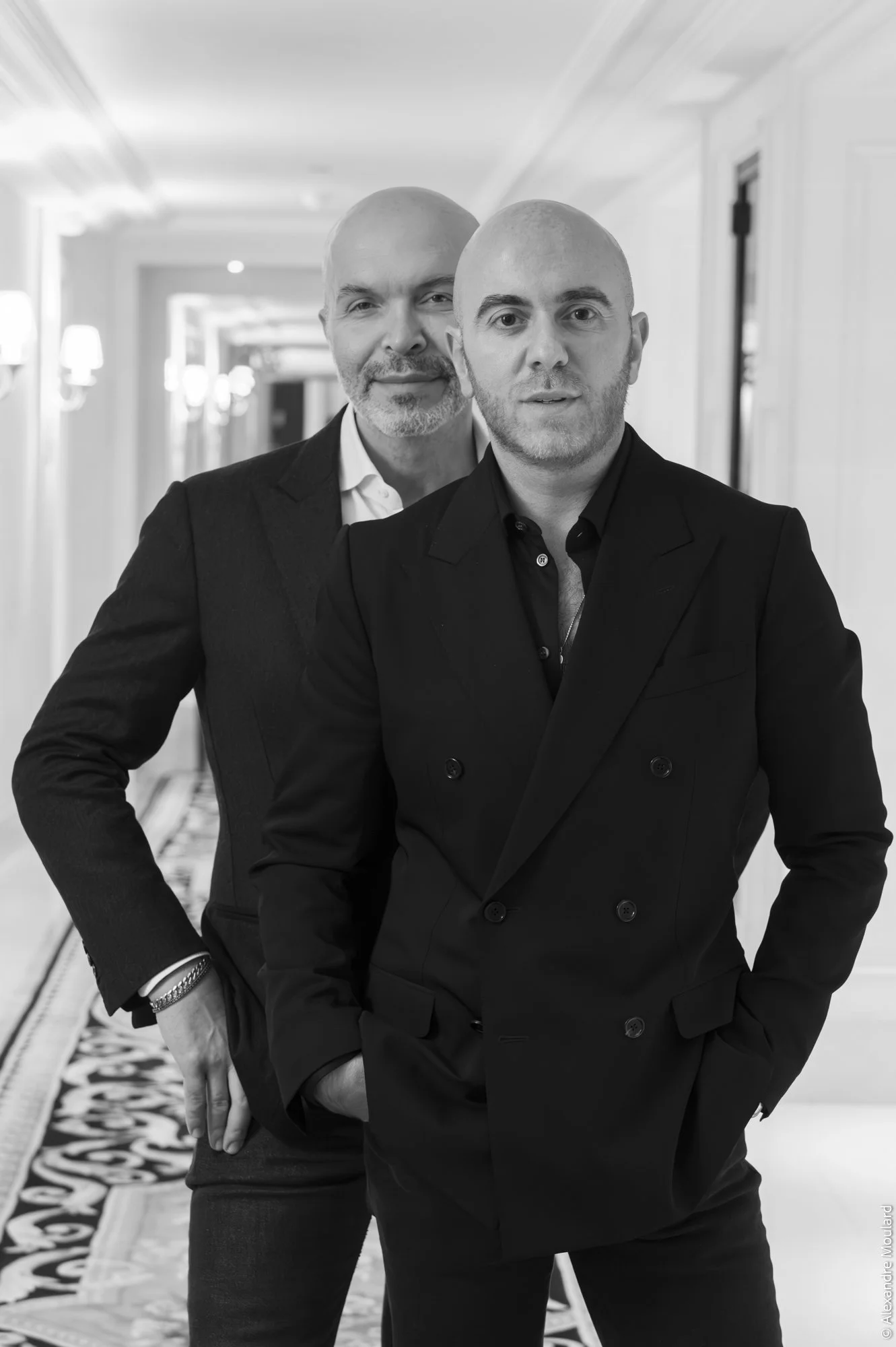 Deux hommes en costume debout dans un couloir élégant, l'un en avant et l'autre derrière, tous deux avec la main dans leur poche.