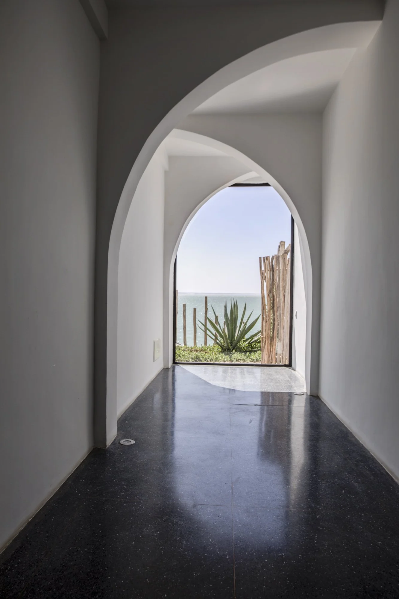 Vue d'un couloir blanc avec une arche et une porte vitrée donnant sur la mer, avec une plante grasse et un panneau en bois à l'extérieur.