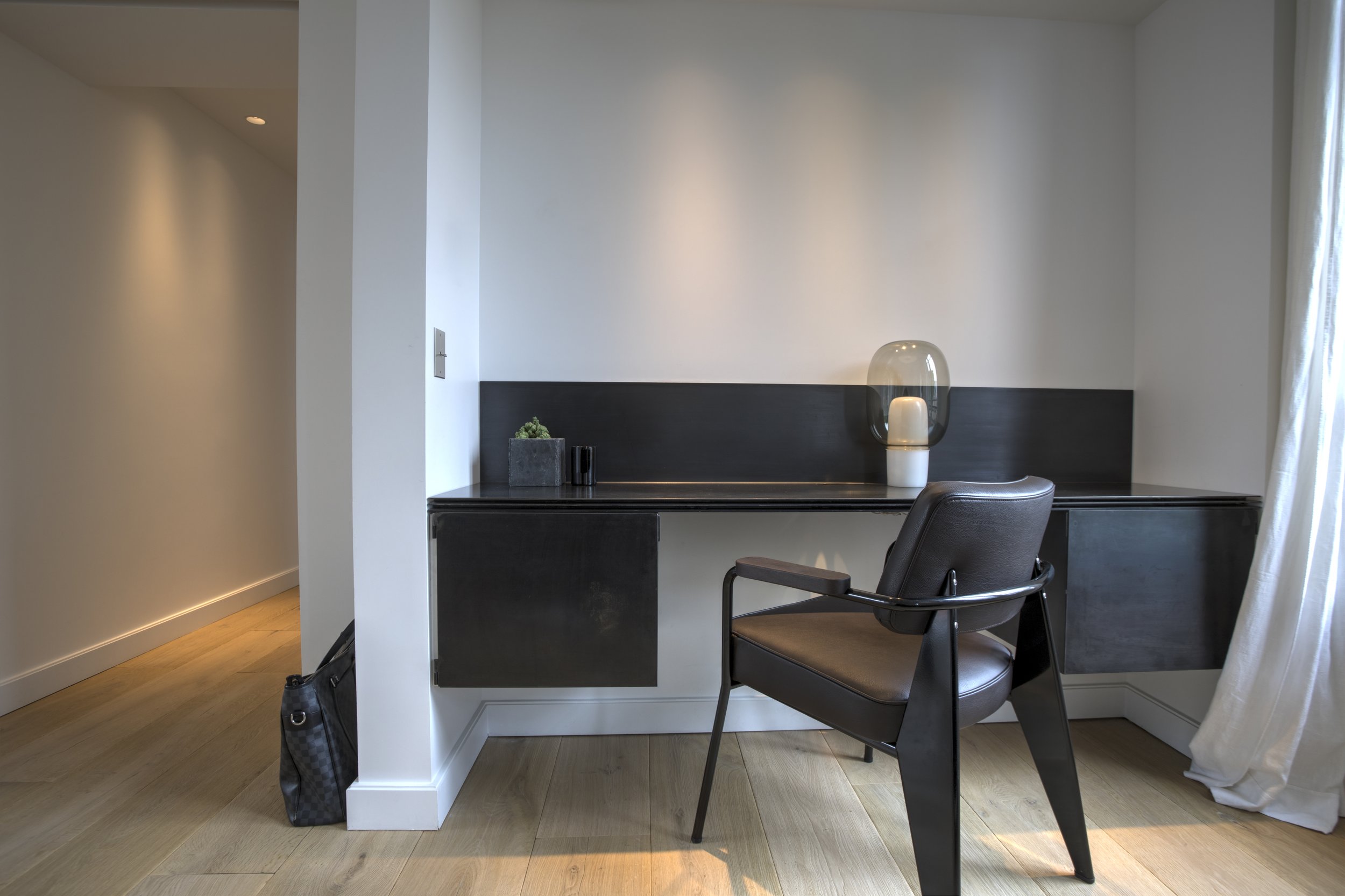 Espace de travail avec un bureau noir, une chaise en cuir, une décoration minimaliste avec un vase en verre et une plante verte, dans une pièce lumineuse avec un rideau blanc.