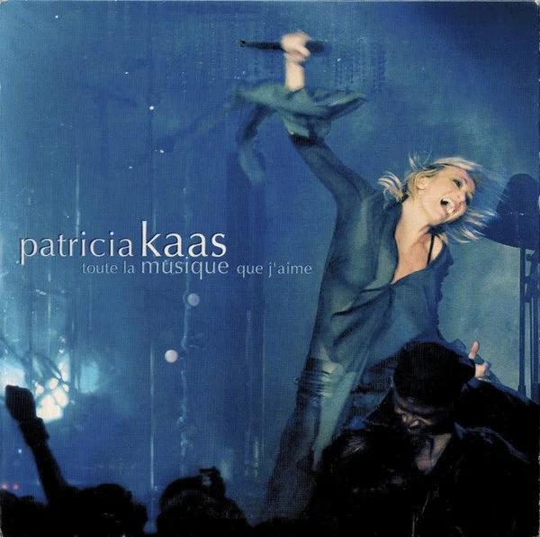 Une femme chante en performante, portant une chemise en jean et tenant un microphone, sur une scène bleue, avec le texte "Patricia Kaas toute la musique que j'aime".