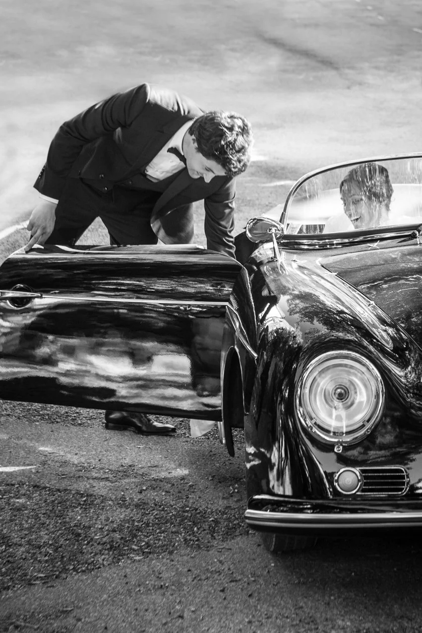 Deux hommes habillés en costume noir, dont un en train de parler ou de regarder quelque chose dans une voiture de sport noire, une Porsche classique.