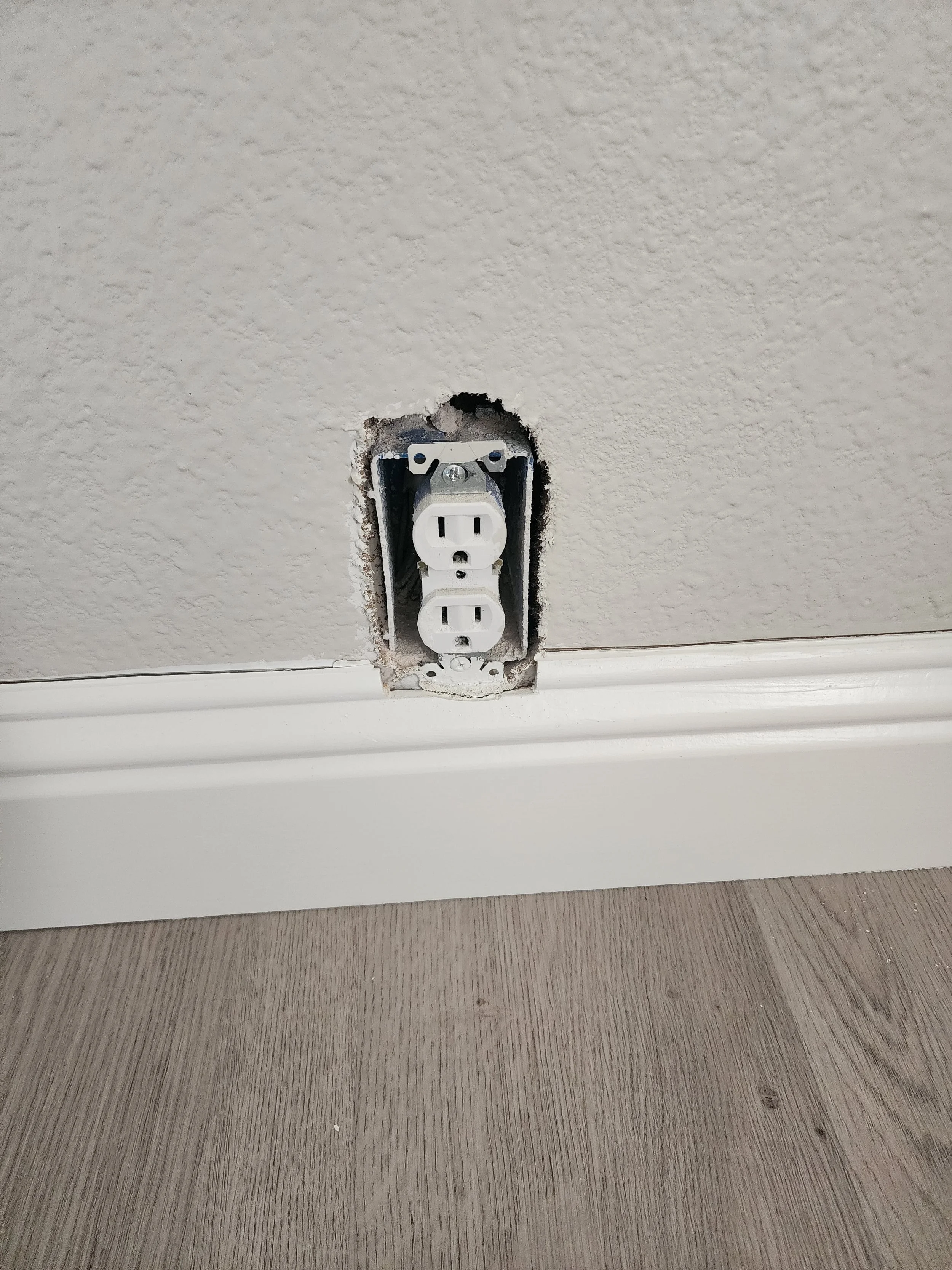 Outlet with a rough, oversized cut-out in the baseboard — functional but unfinished.