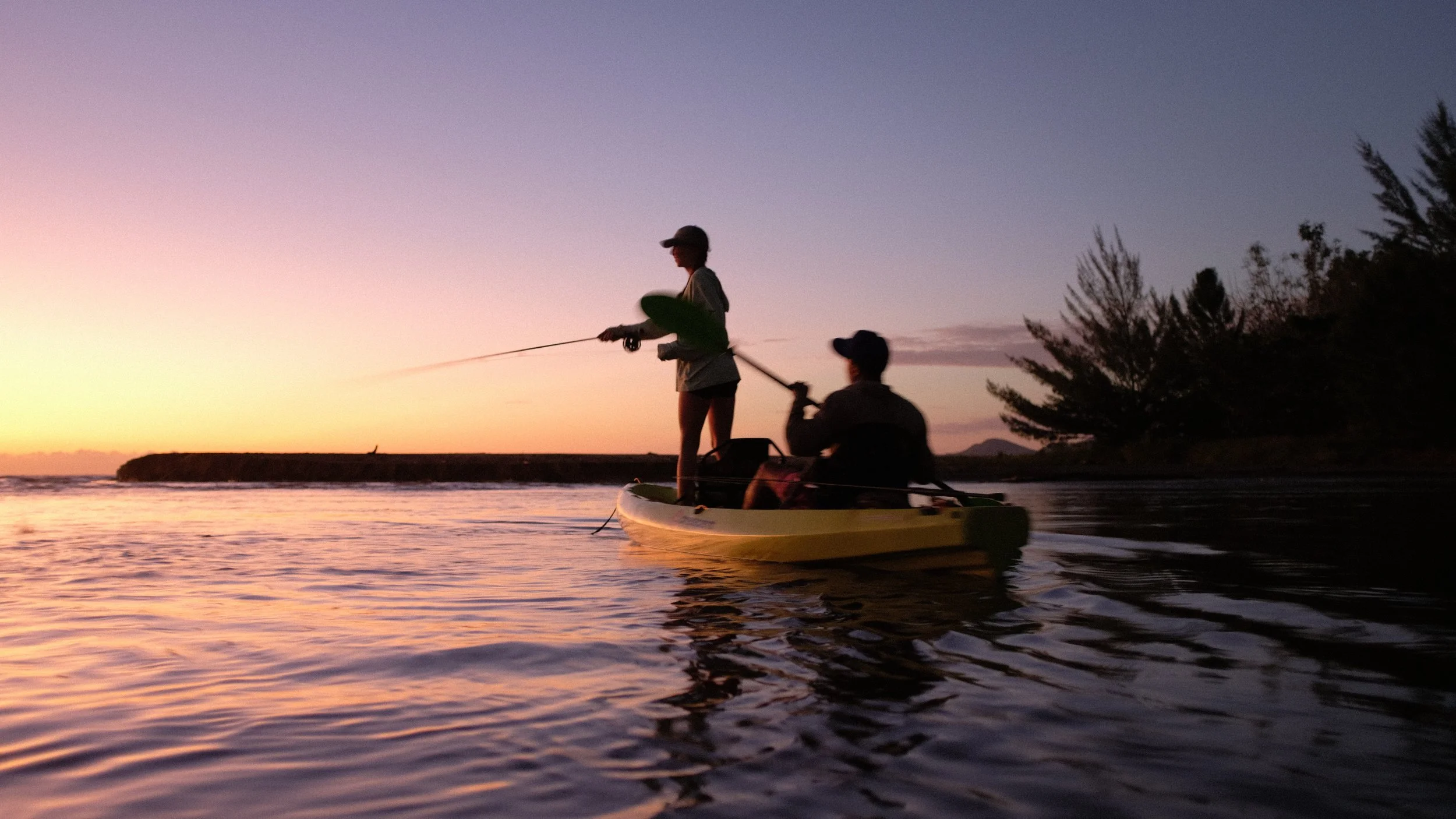 Two people fishing from a kayak on calm water during sunset, with trees and a distant landmass in the background.