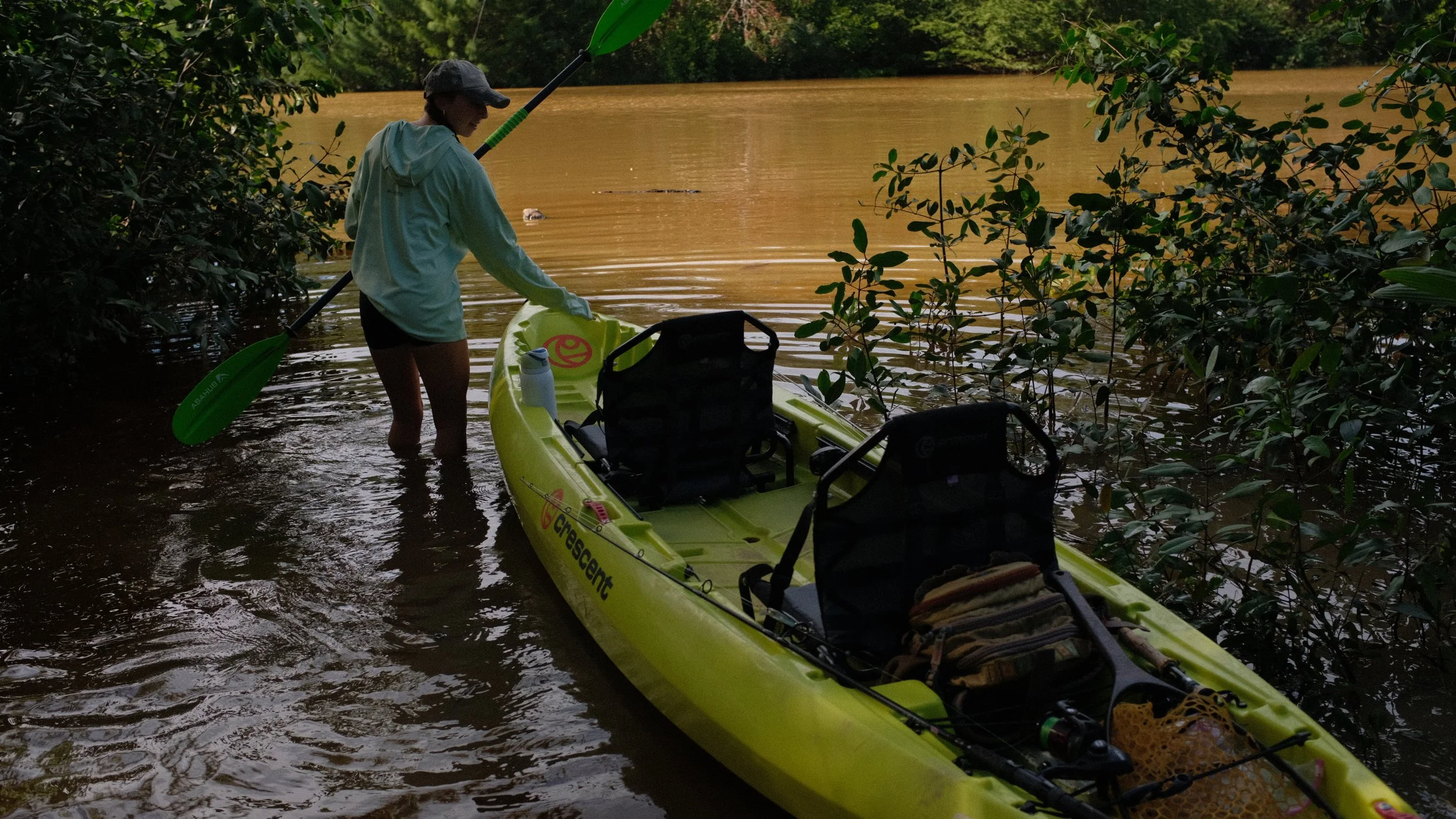 Person launching a yellow kayak into a river surrounded by trees with muddy water.