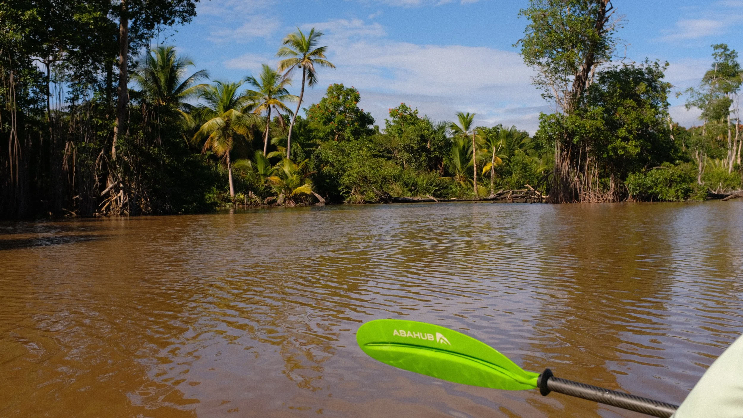 View from a kayak on a brown river with lush green tropical trees and palms along the riverbank, under a partly cloudy sky.