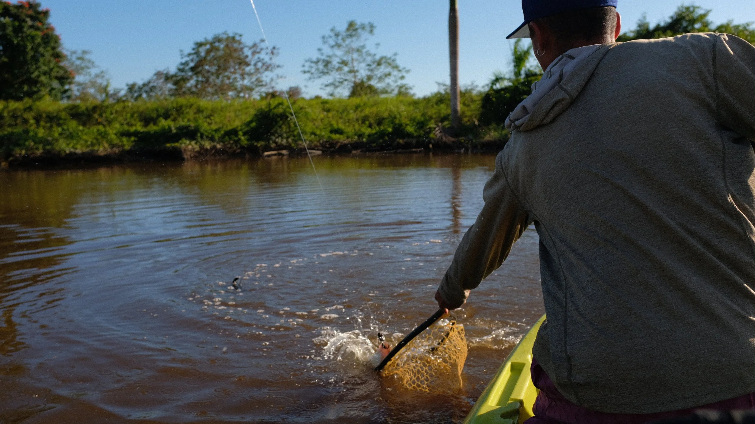 Person in a gray hoodie fishing in a river, with trees and bushes in the background, during daytime.