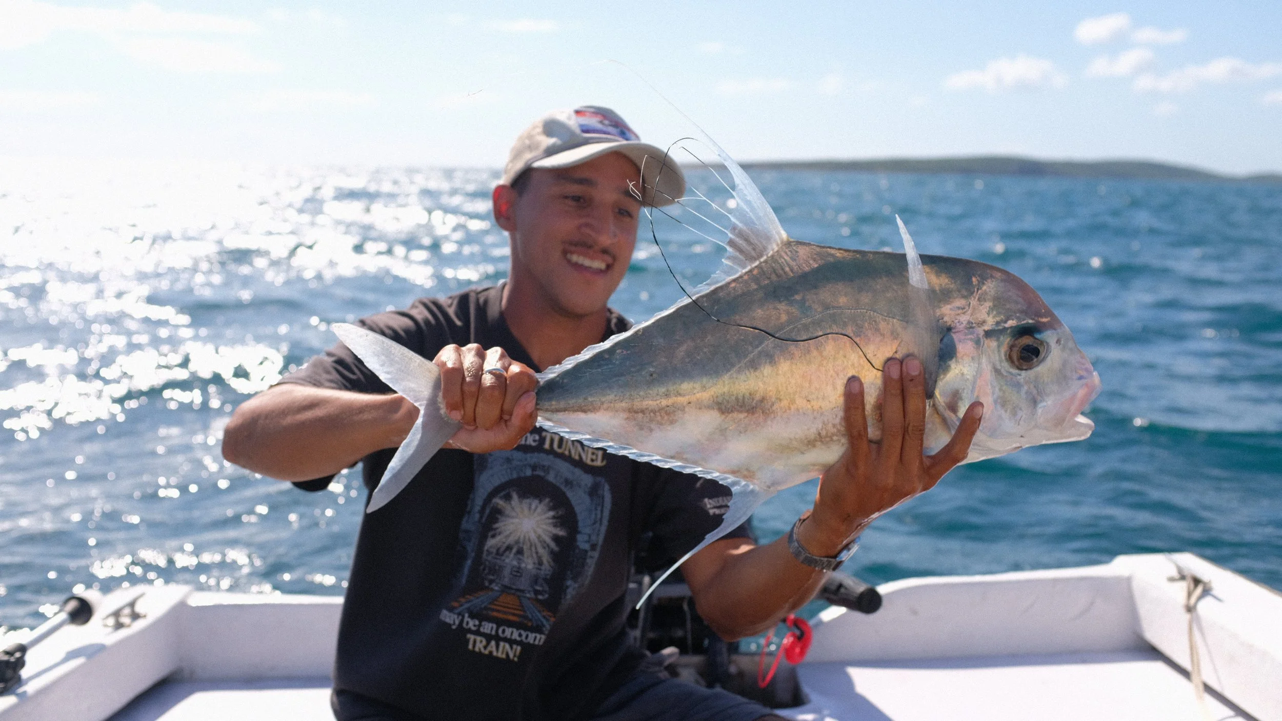 A man on a boat holding a large fish with a fishing line hanging from its mouth, smiling at the camera, with the ocean and sky in the background.