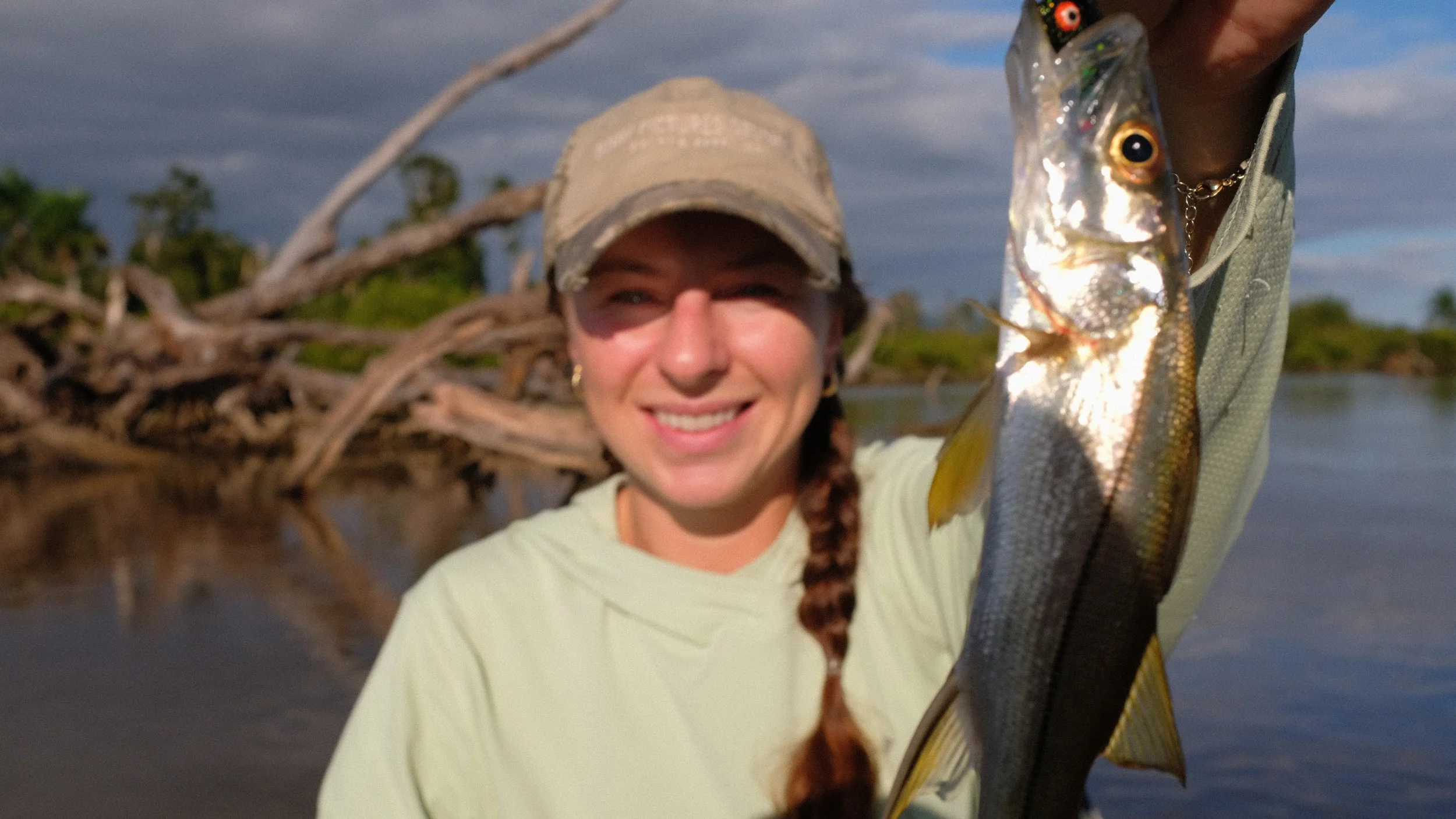 A smiling woman holding up a fish she caught, standing near a river with fallen trees in the background.