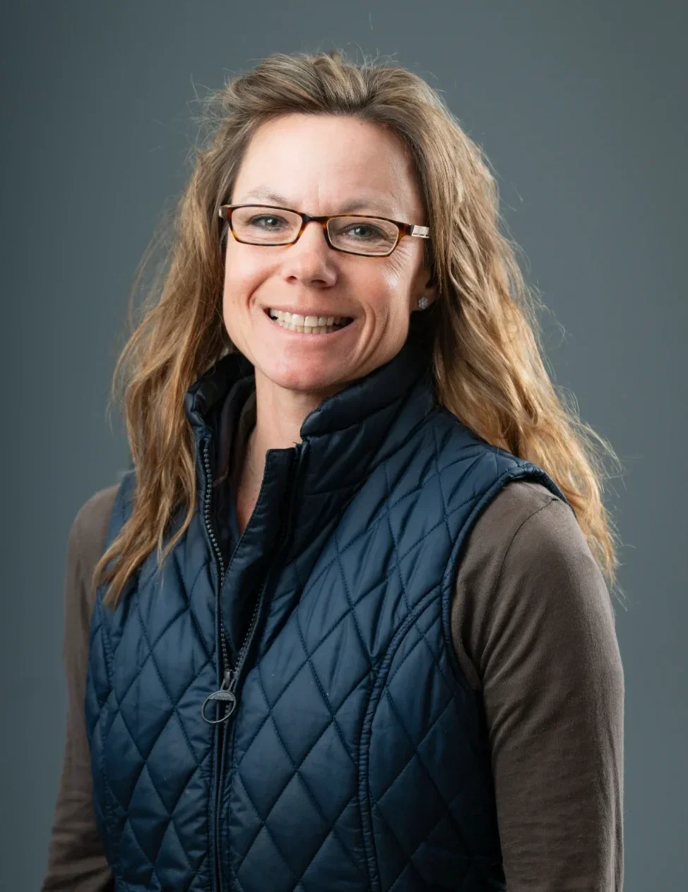 A woman with wavy light brown hair, wearing glasses, a brown long-sleeve shirt, and a navy blue quilted vest, smiling at the camera against a gray background.