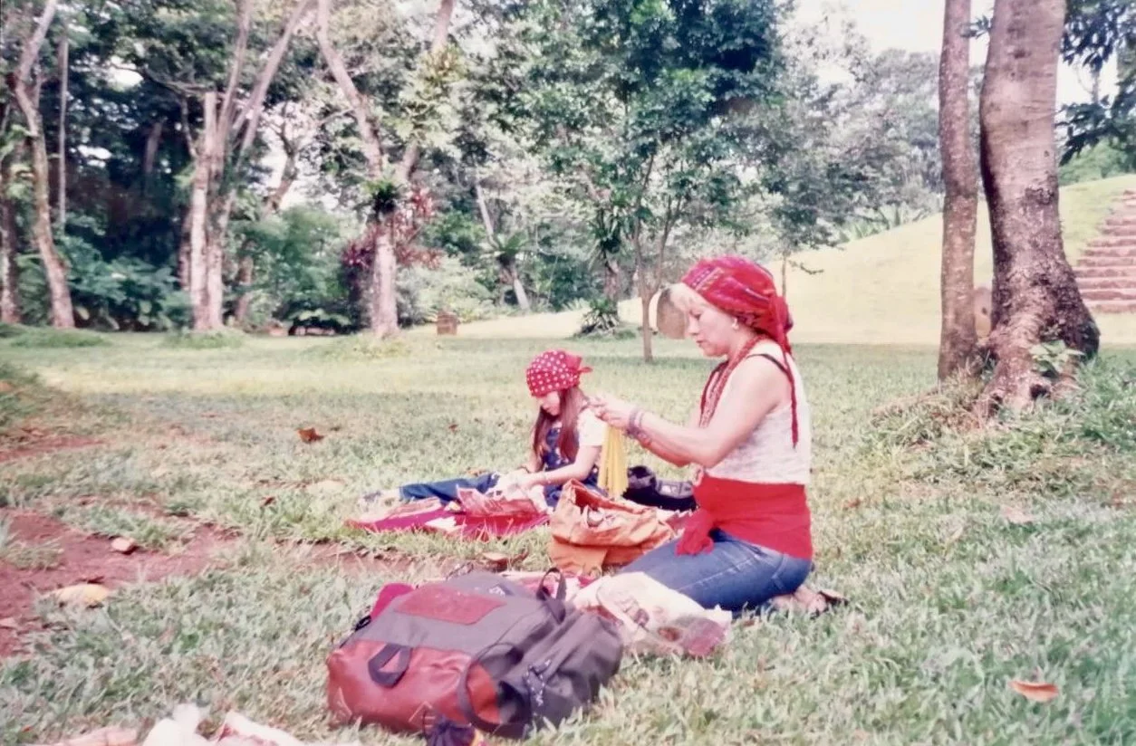 A woman and a girl sitting on the grass in a park, surrounded by trees, with backpacks and outdoor gear, enjoying a picnic or outdoor activity.