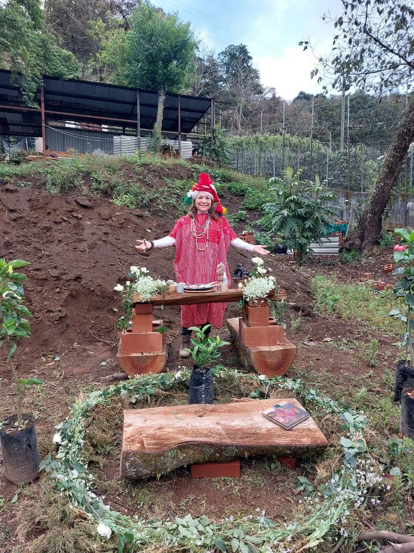 A woman dressed in a red and pink striped outfit with a red hat, standing outdoors in a garden, smiling and gesturing with her arms open. There is a rustic wooden table in front of her decorated with flowers, and plants around the garden.
