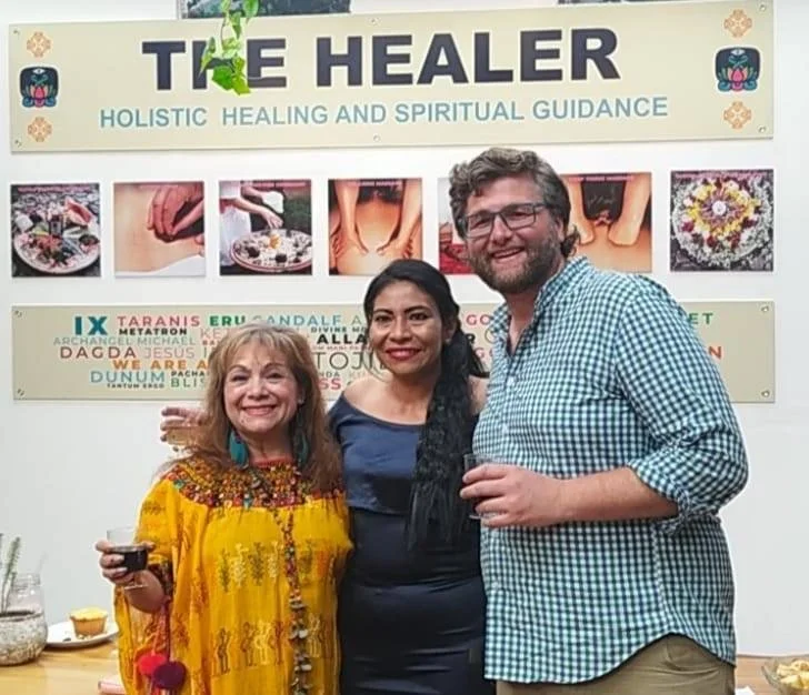 Three people smiling and posing in front of a sign that reads 'The Healer: Holistic Healing and Spiritual Guidance' with various images and text about healing methods behind them.