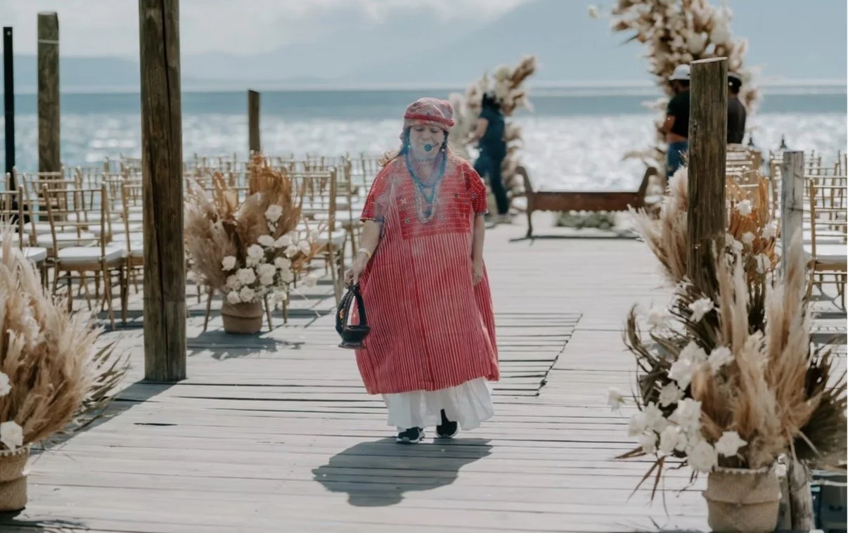 A woman walking down a wooden aisle decorated with white flowers and pampas grass at an outdoor beach setting, with chairs on either side and the ocean in the background. She is dressed in a colorful, bohemian-style outfit with a hat and carries a small bag.