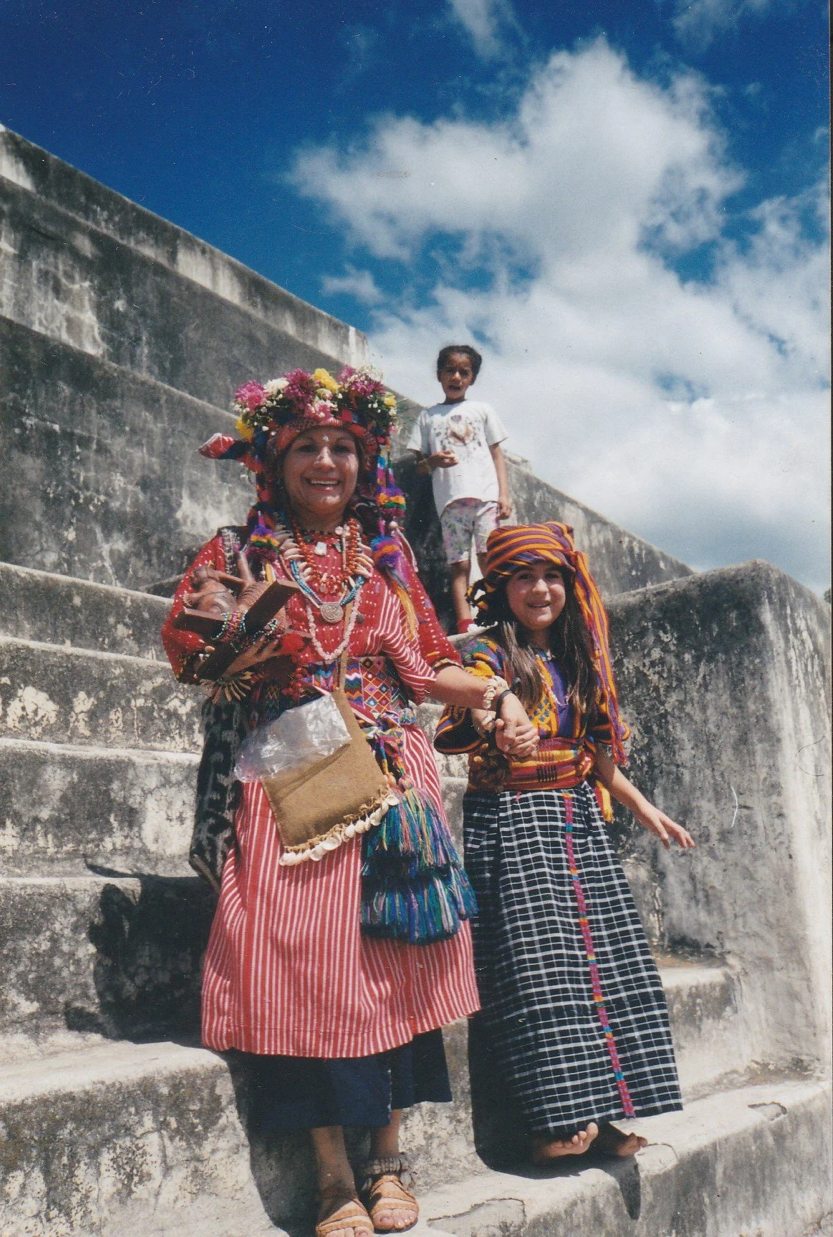 A woman dressed in traditional colorful attire with a flower crown and jewelry, holding hands with a young girl in similar traditional clothing, both standing on stone stairs outdoors with a blue sky and clouds in the background. A child is standing behind them on the stairs.
