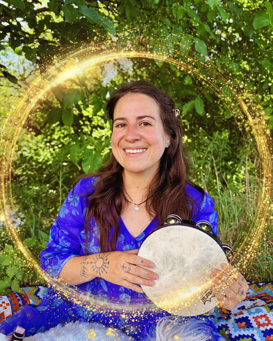 A woman with long brown hair in a blue patterned outfit sitting outdoors among green foliage, holding a tambourine and smiling at the camera, with golden light effects surrounding her.