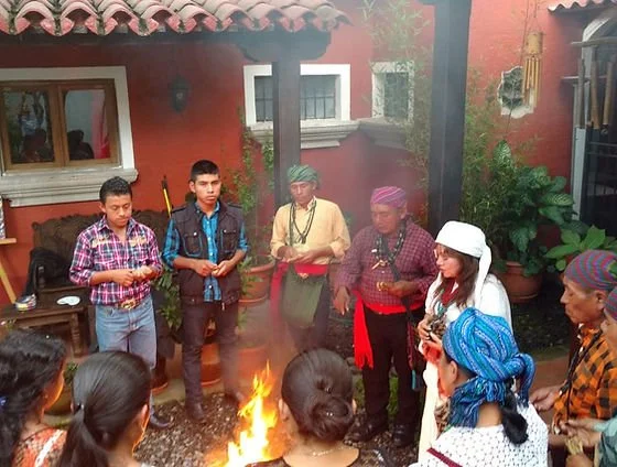 Group of people gathered around a small fire outdoors, participating in a cultural or ceremonial activity, with traditional clothing and plants in the background.
