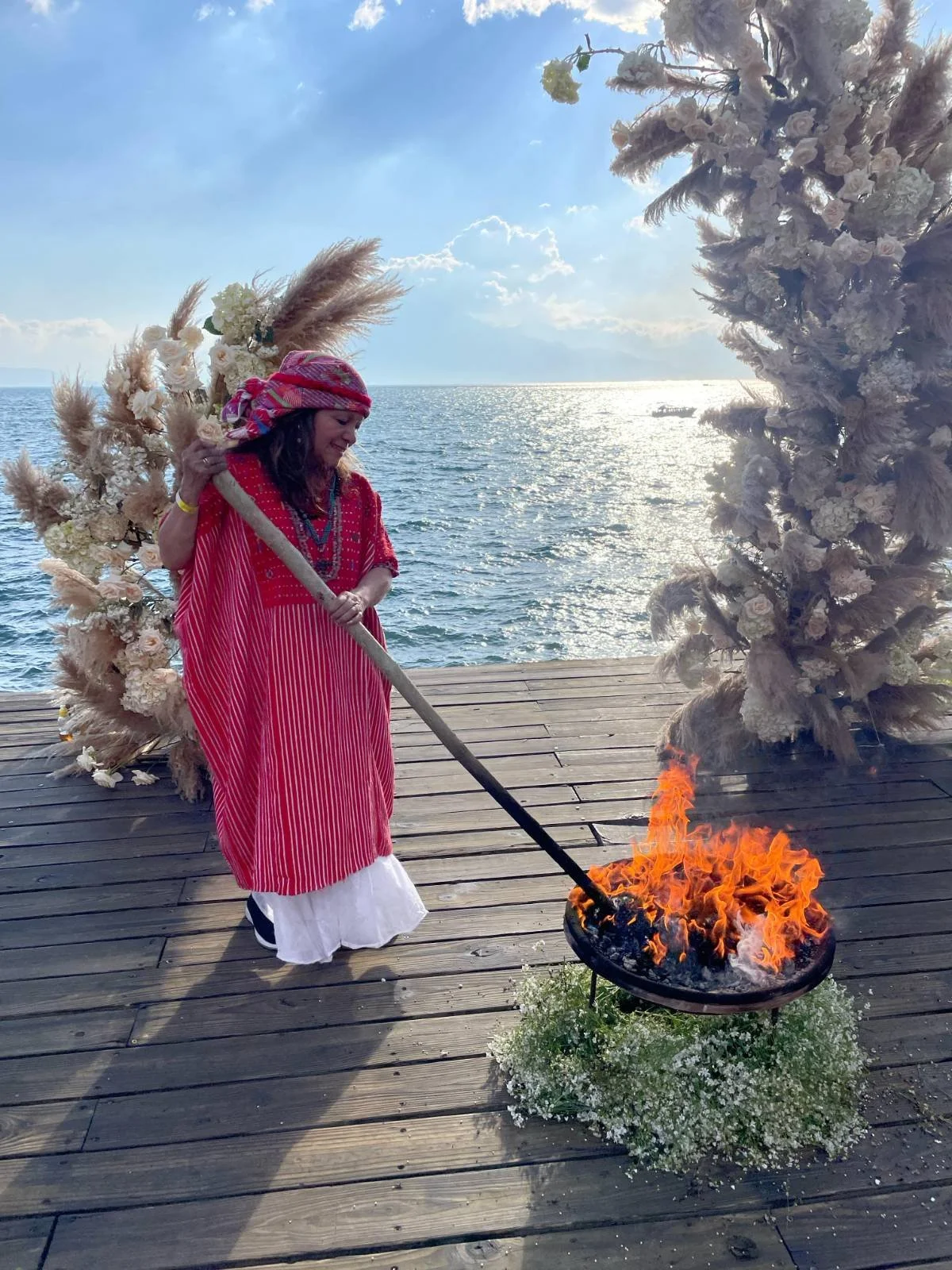 A woman in traditional clothing stands on a wooden dock by the water, holding a long stick with a flaming fire pit at the end, surrounded by floral arrangements and a backdrop of the ocean and sky.