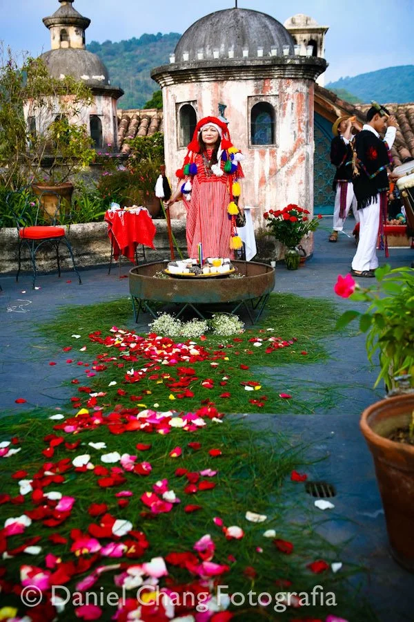 Person in colorful traditional attire standing on a terrace decorated with scattered rose petals, with a small altar and flowers, in front of a historic building with domed towers, accompanied by musicians in traditional dress.