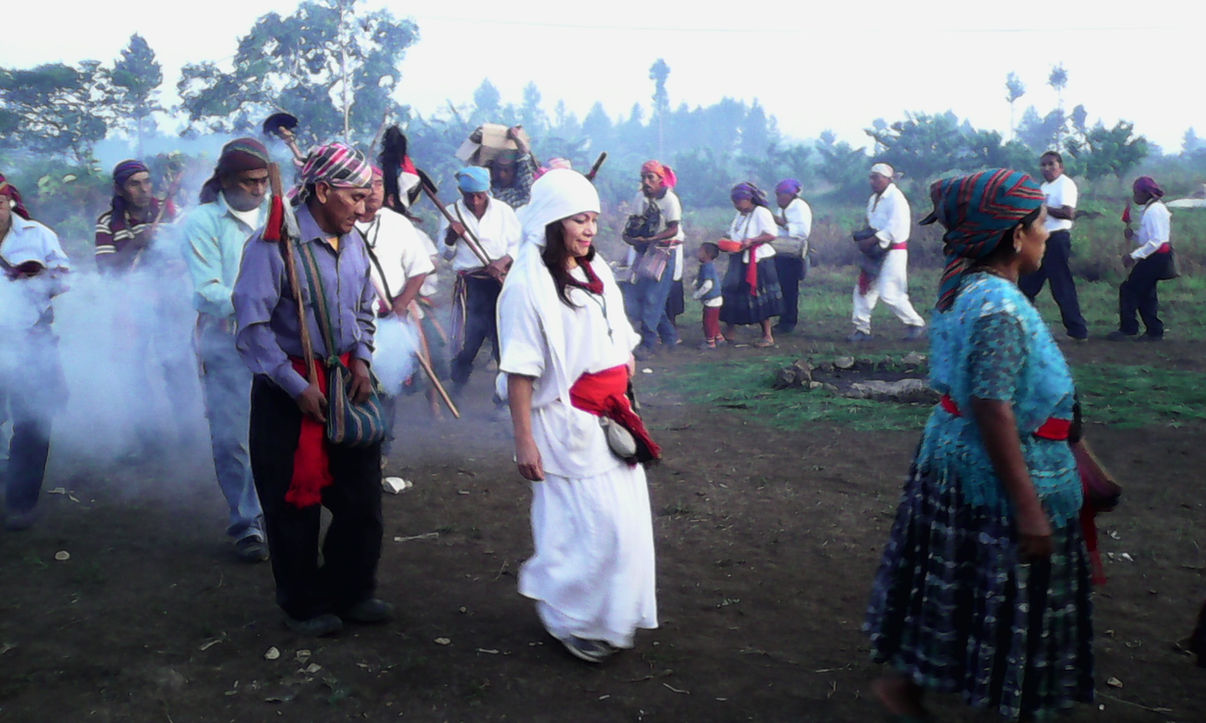Group of people dressed in traditional clothing walking outdoors in a rural area with trees in the background.