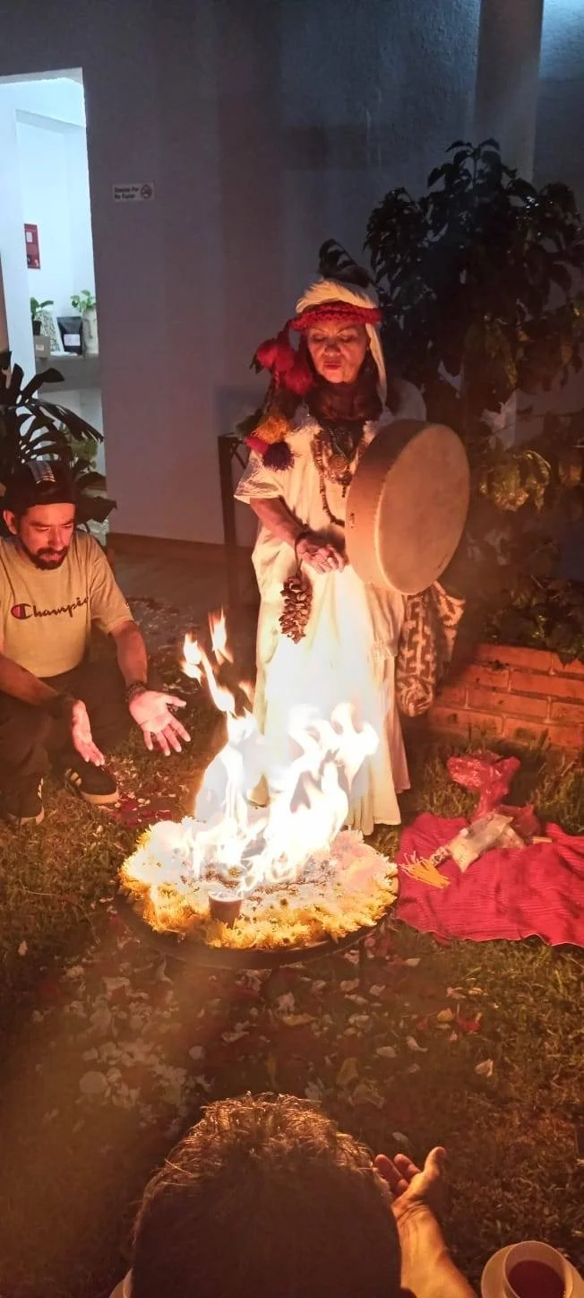 Person dressed in traditional indigenous attire standing near a fire with flames, holding a drum, surrounded by other individuals, outdoors during evening.
